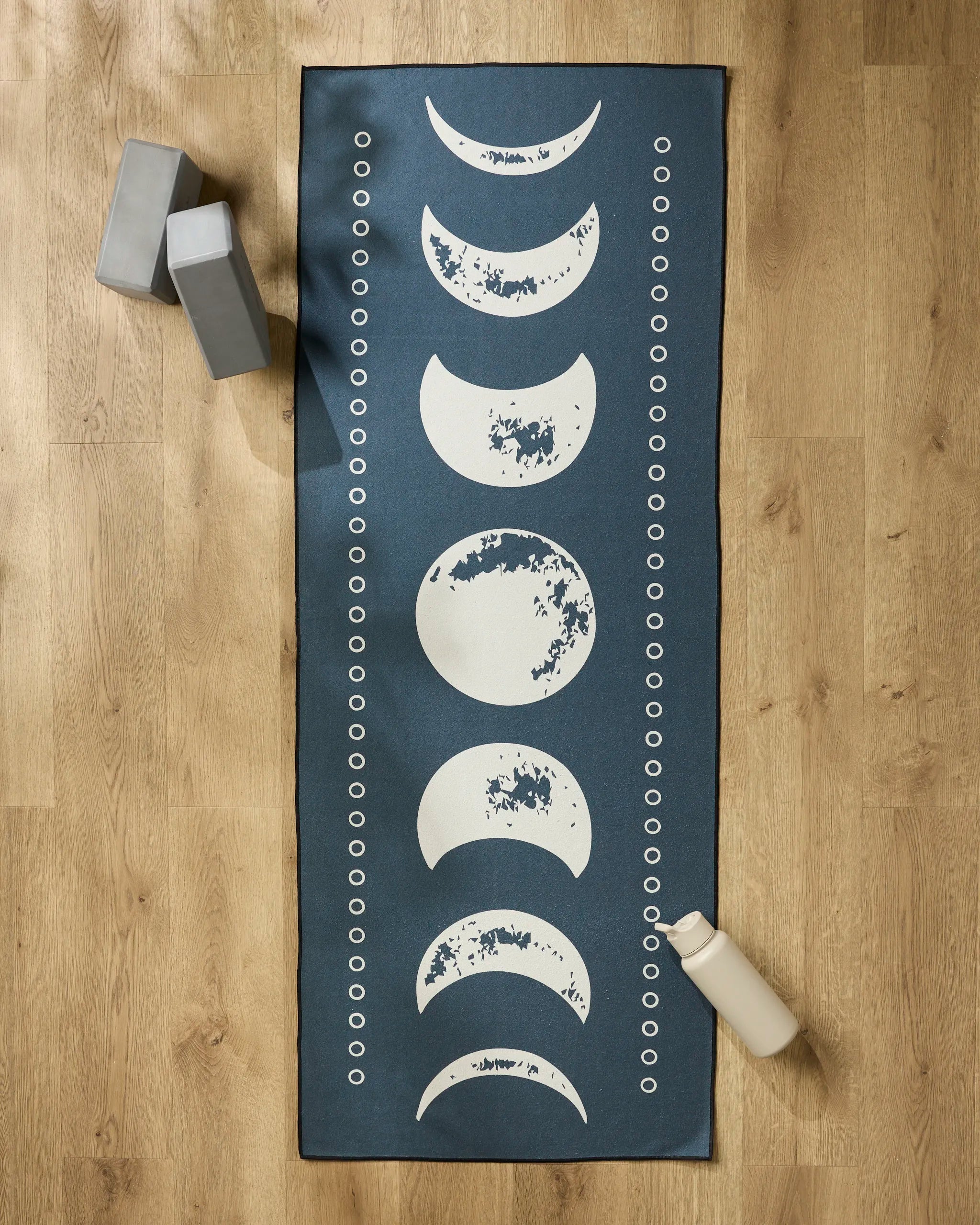 The Ashley Celena Lunar Phases blue yoga mat, featuring white moon phase designs and small white circles, is laid on a wooden floor with a slip-resistant yoga towel, two gray blocks, and a beige water bottle nearby.