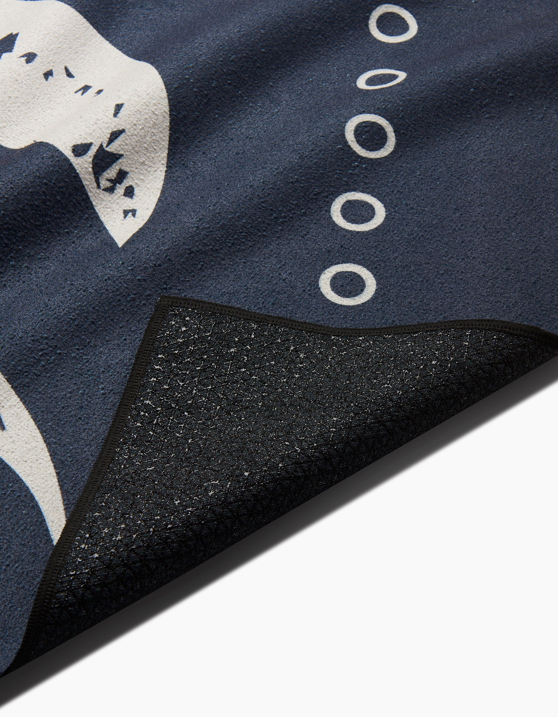 A close-up of Ashley Celena’s Lunar Phases yoga towel in dark blue with white abstract and circular designs, showing its textured black recycled underside partially folded over.