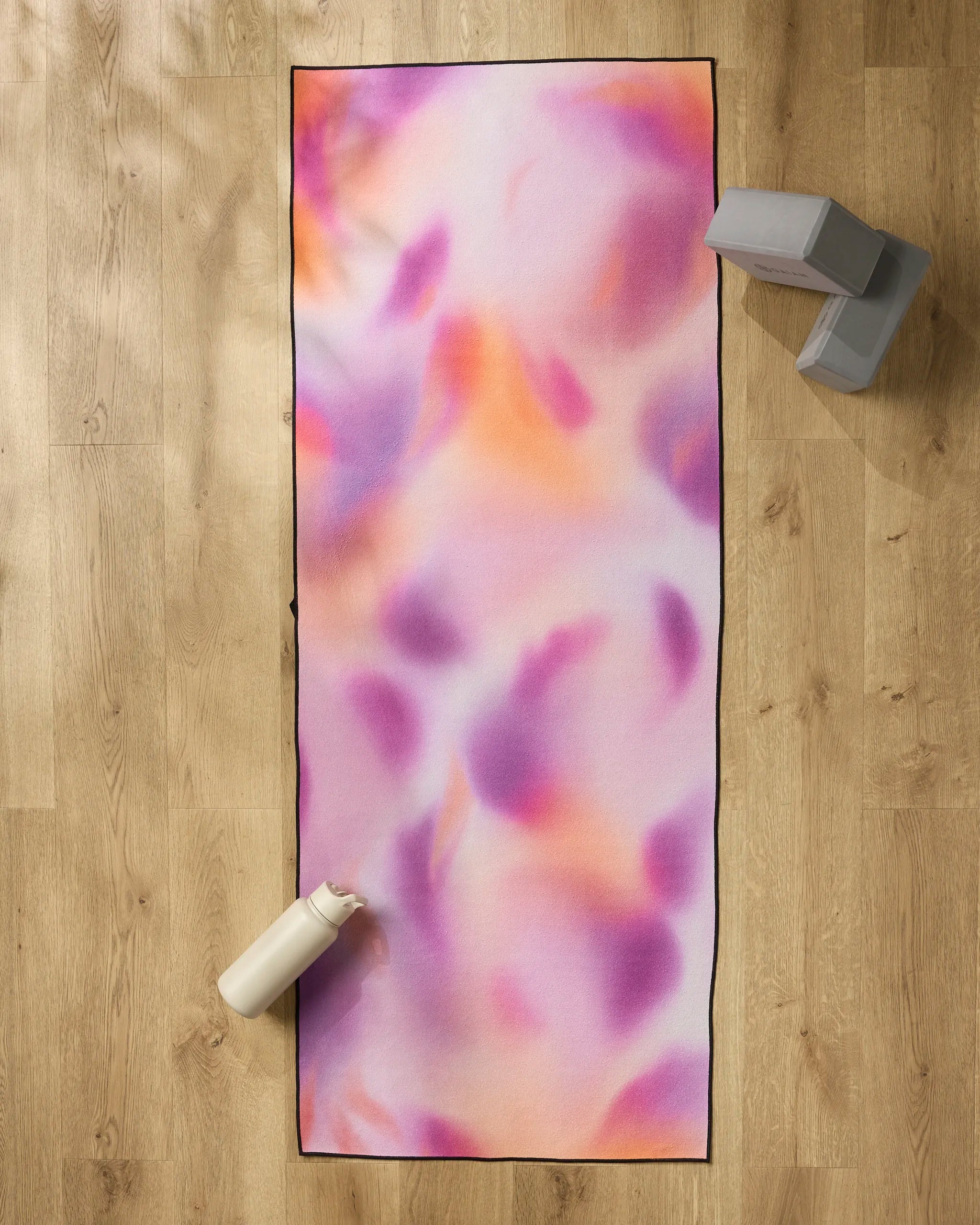 The GEOMETRY Magenta yoga mat, featuring pink, orange, and purple swirls, rests on a wooden floor. A white water bottle sits at the bottom left corner with two gray yoga blocks and a non-slip towel at the top right.
