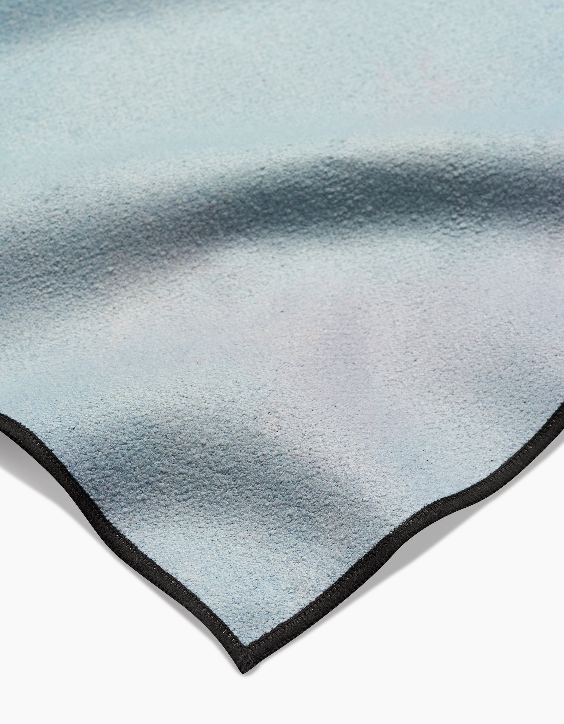 A detailed view of Stefanie Bales’ Magic in the Sky yoga towel, crafted from recycled materials in a soft light blue hue, showcasing a black smooth-edged border, gentle folds, and a subtle color gradient.