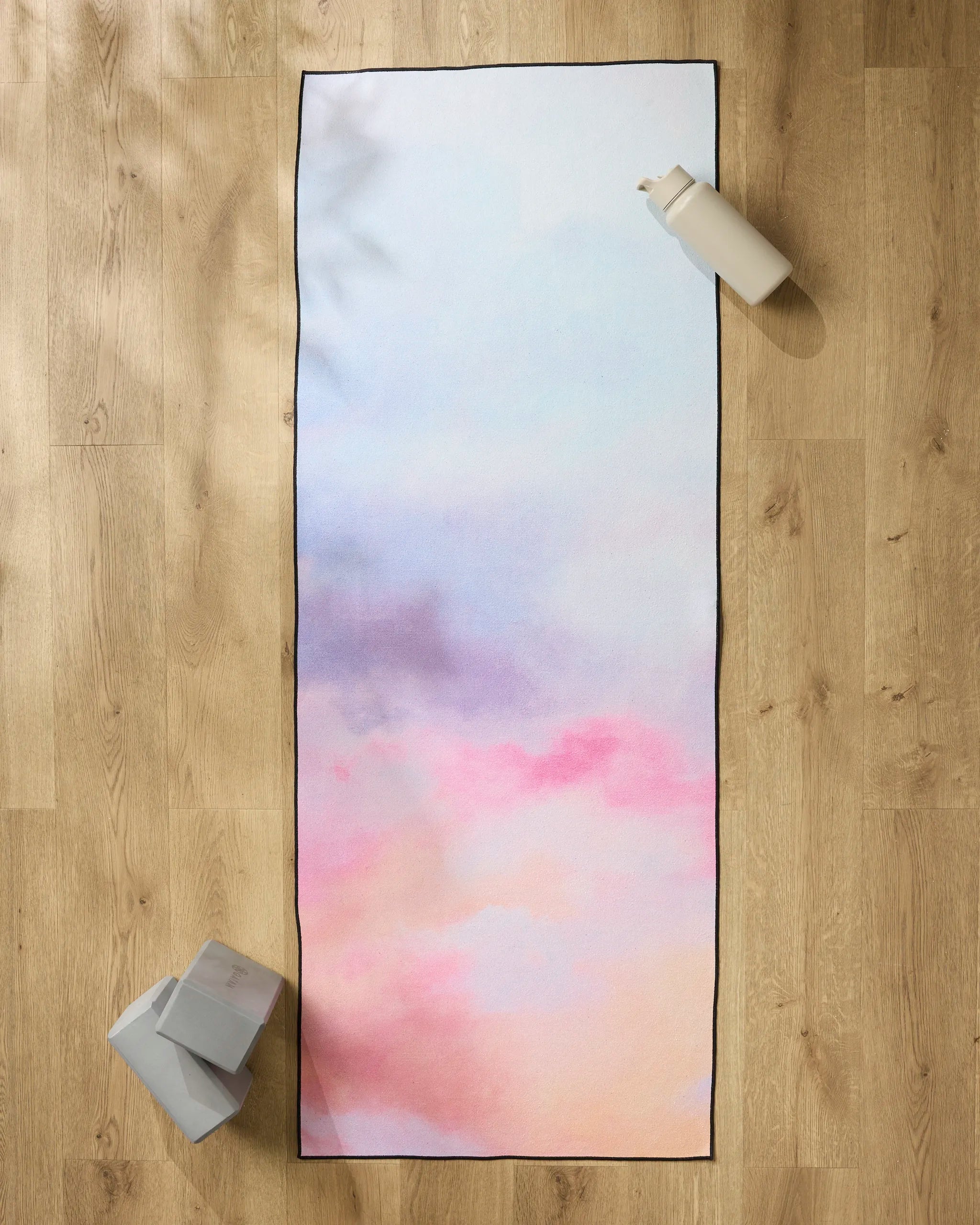 The "Magic in the Sky" yoga mat by Stefanie Bales, featuring pastel pink, purple, and blue hues, rests on a wooden floor with a white water bottle, two gray yoga blocks, and a silicone grip towel nearby.