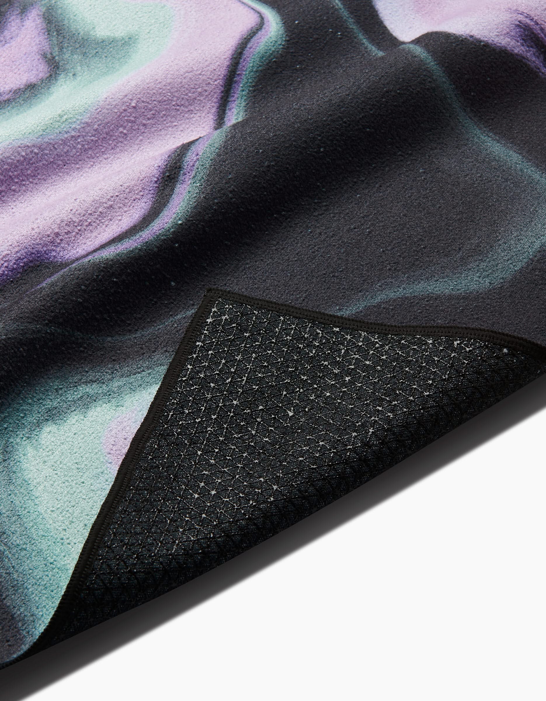 A close-up of the GEOMETRY Marble yoga mat shows its textured, non-slip black underside and a smooth top with a purple, green, and black marbled design. One corner is folded over to reveal both sides—perfect with a slip-resistant yoga towel.