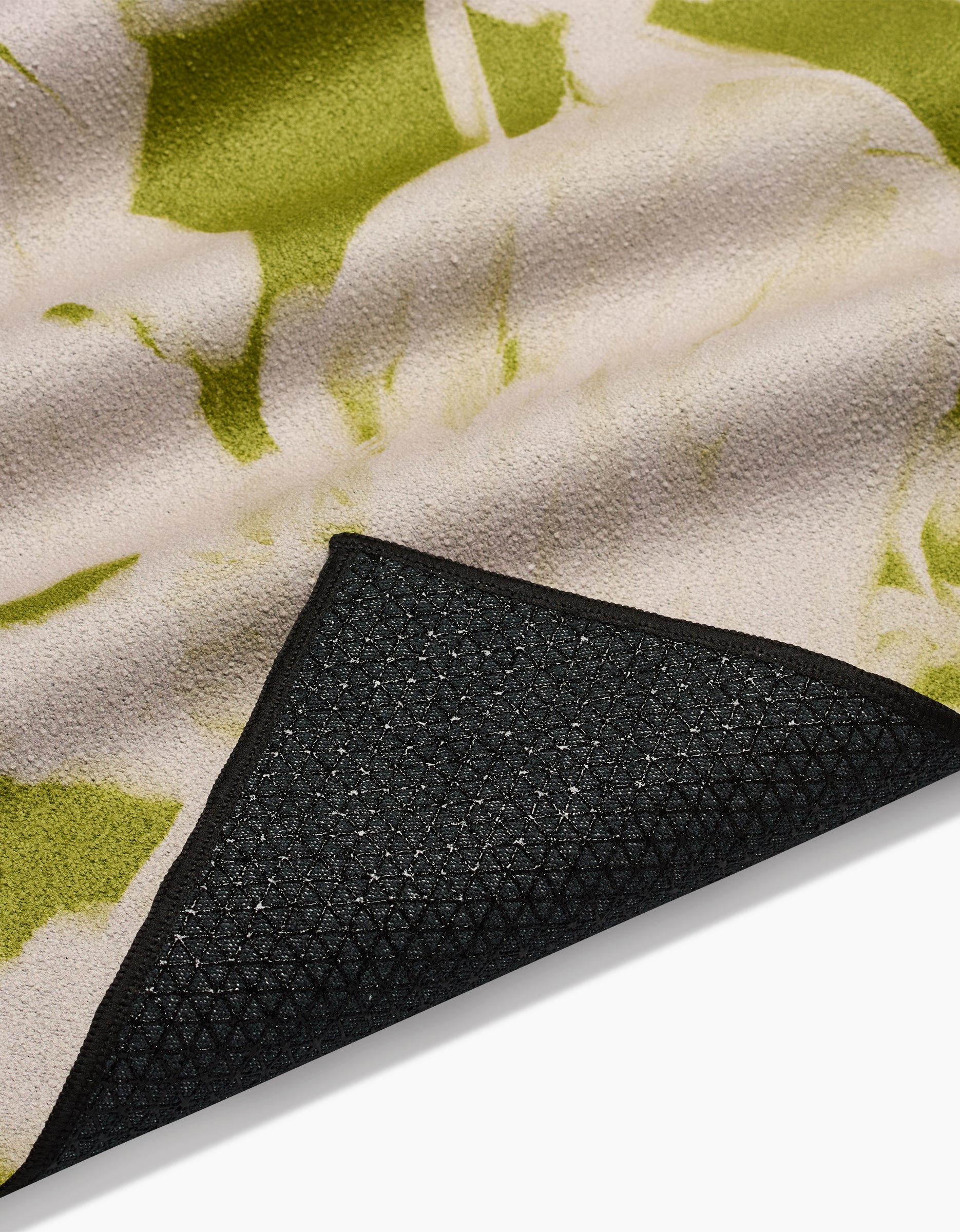 A close-up of GEOMETRYs Meadow Mist yoga towel shows its green and beige abstract pattern and black textured silicone grip non-slip backing, partially folded to reveal the underside—crafted from recycled materials.