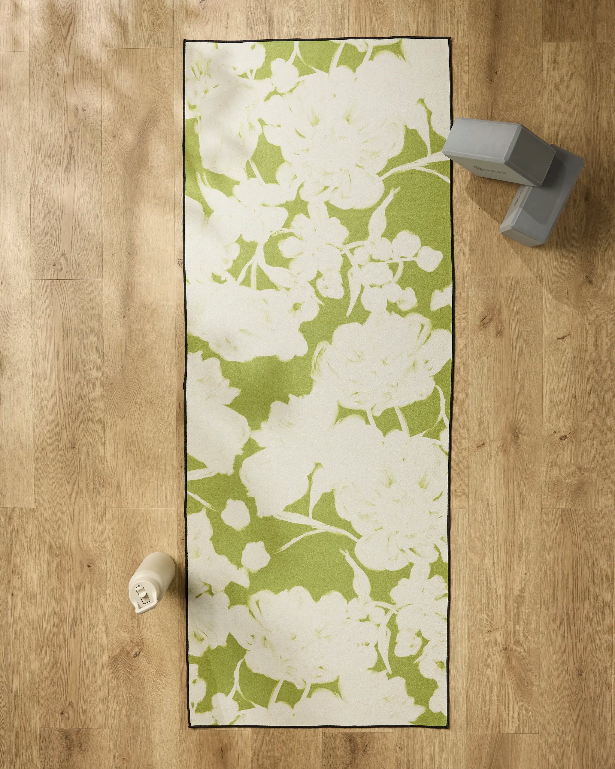 The Meadow Mist yoga mat by GEOMETRY, featuring a green and white floral design, is spread on a wooden floor with a non-slip towel. A water bottle rests at the bottom left and two gray yoga blocks are stacked at the top right.