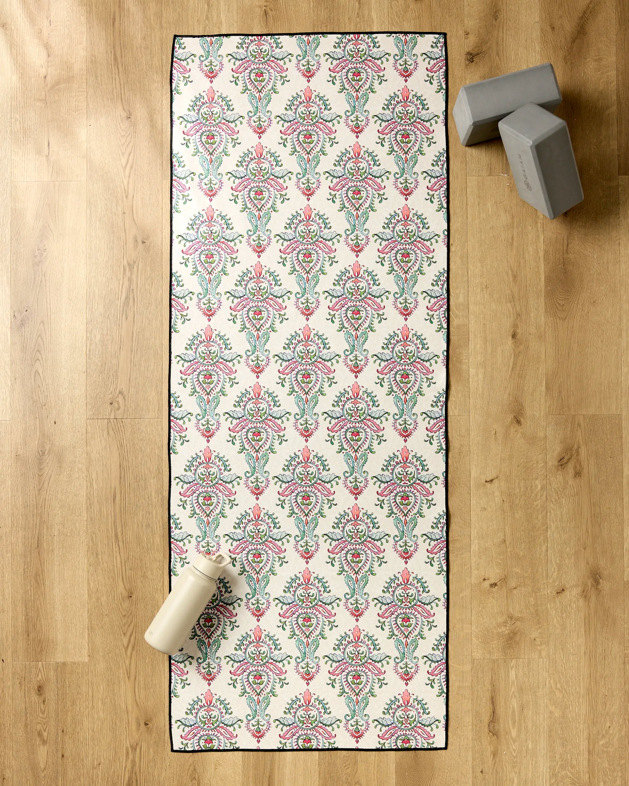 The Memento yoga mat by Maayan Alper-Swan features a pink and green floral pattern on a wooden floor, paired with a non-slip towel, a white water bottle at the bottom left, and two gray blocks stacked top right.