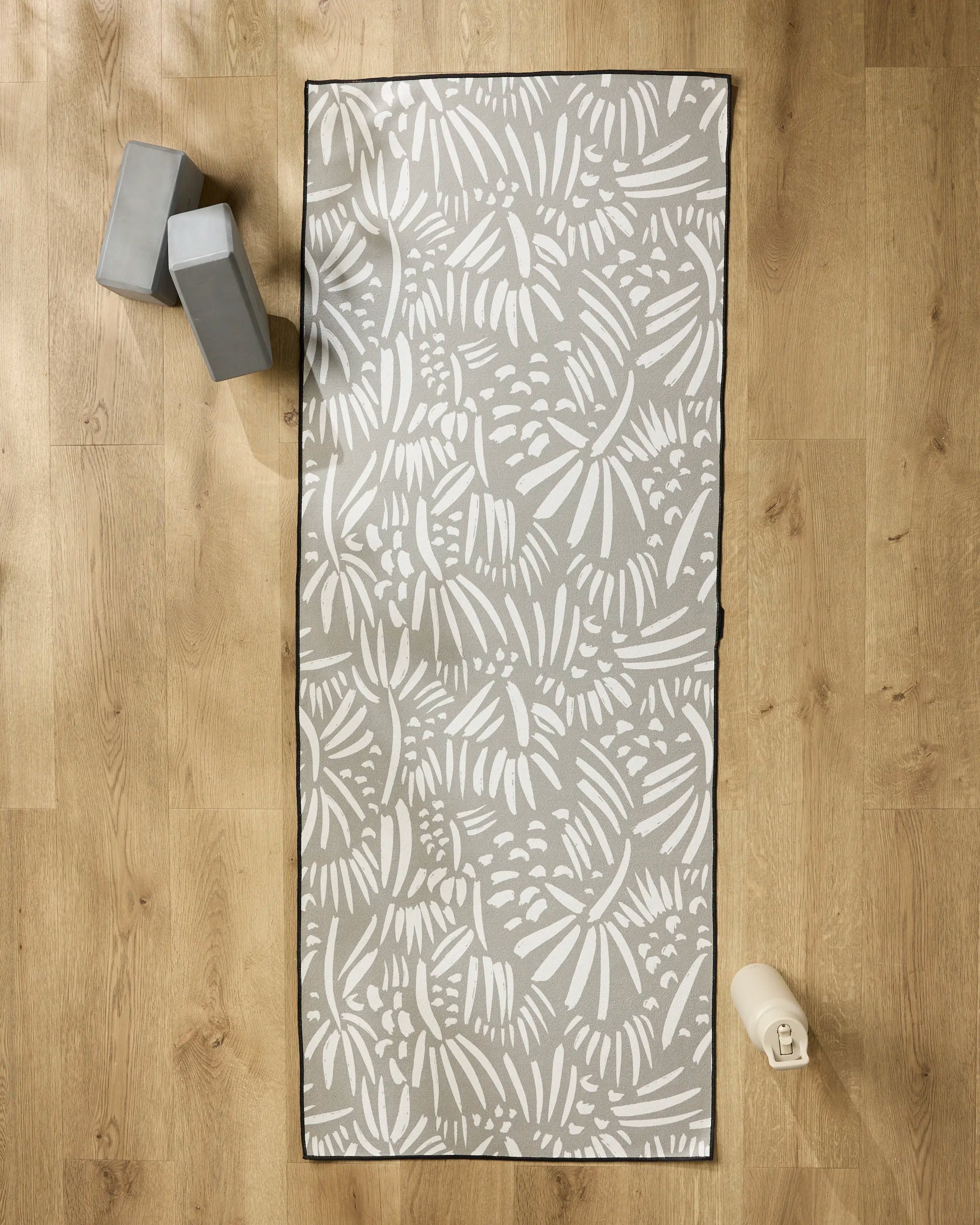The Miami yoga mat by Hufton Studio, featuring white abstract leaf designs on a gray background, is shown on a wooden floor with two gray blocks, a water bottle, and a non-slip towel to enhance your practice.