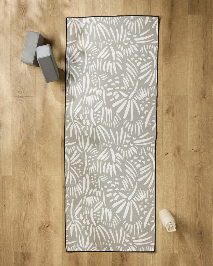 The Miami yoga mat by Hufton Studio, featuring white abstract leaf designs on a gray background, is shown on a wooden floor with two gray blocks, a water bottle, and a non-slip towel to enhance your practice.