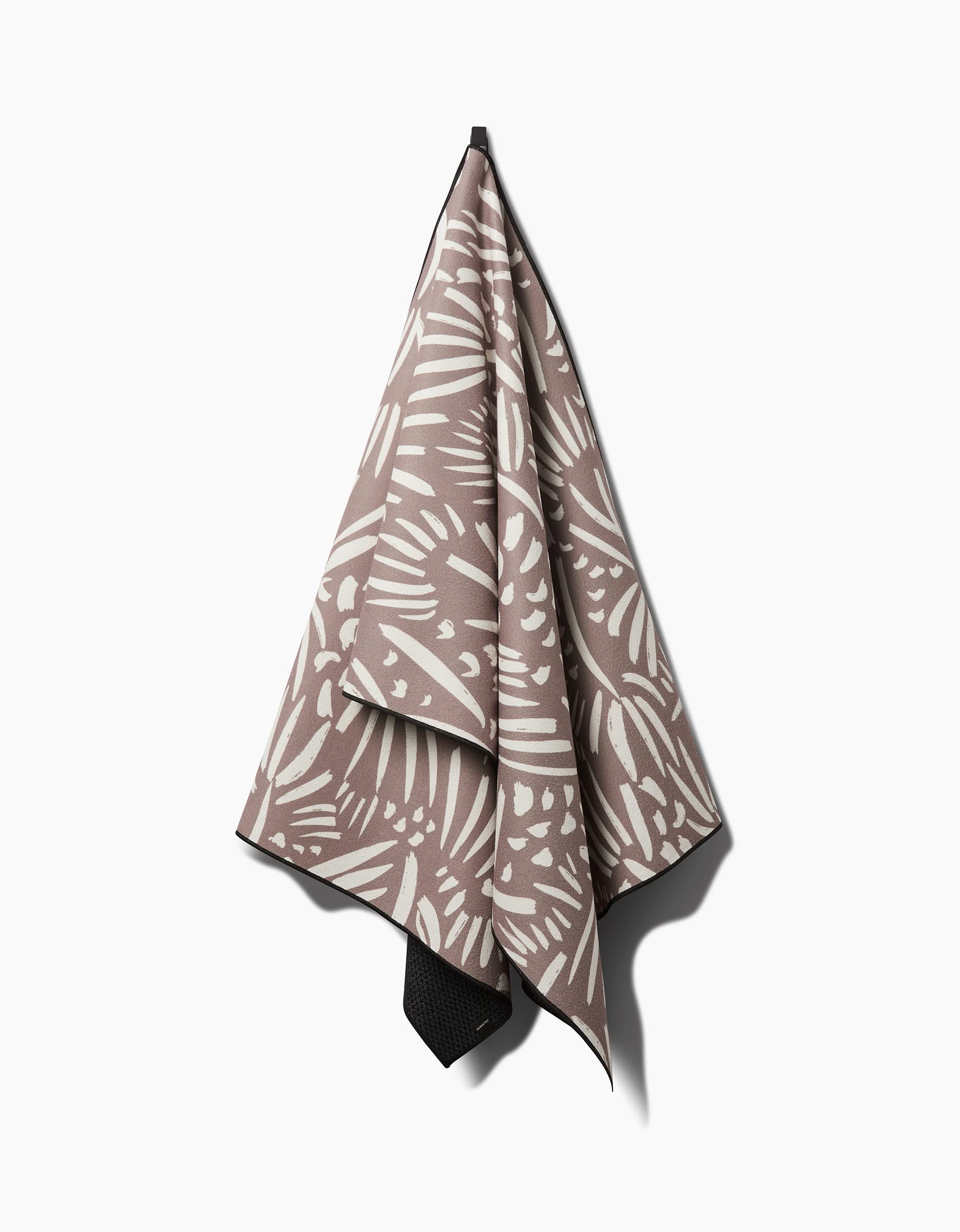 The Miami yoga towel by Hufton Studio features a beige and white abstract leaf pattern and a non-slip design, shown hanging on a hook against a white background.