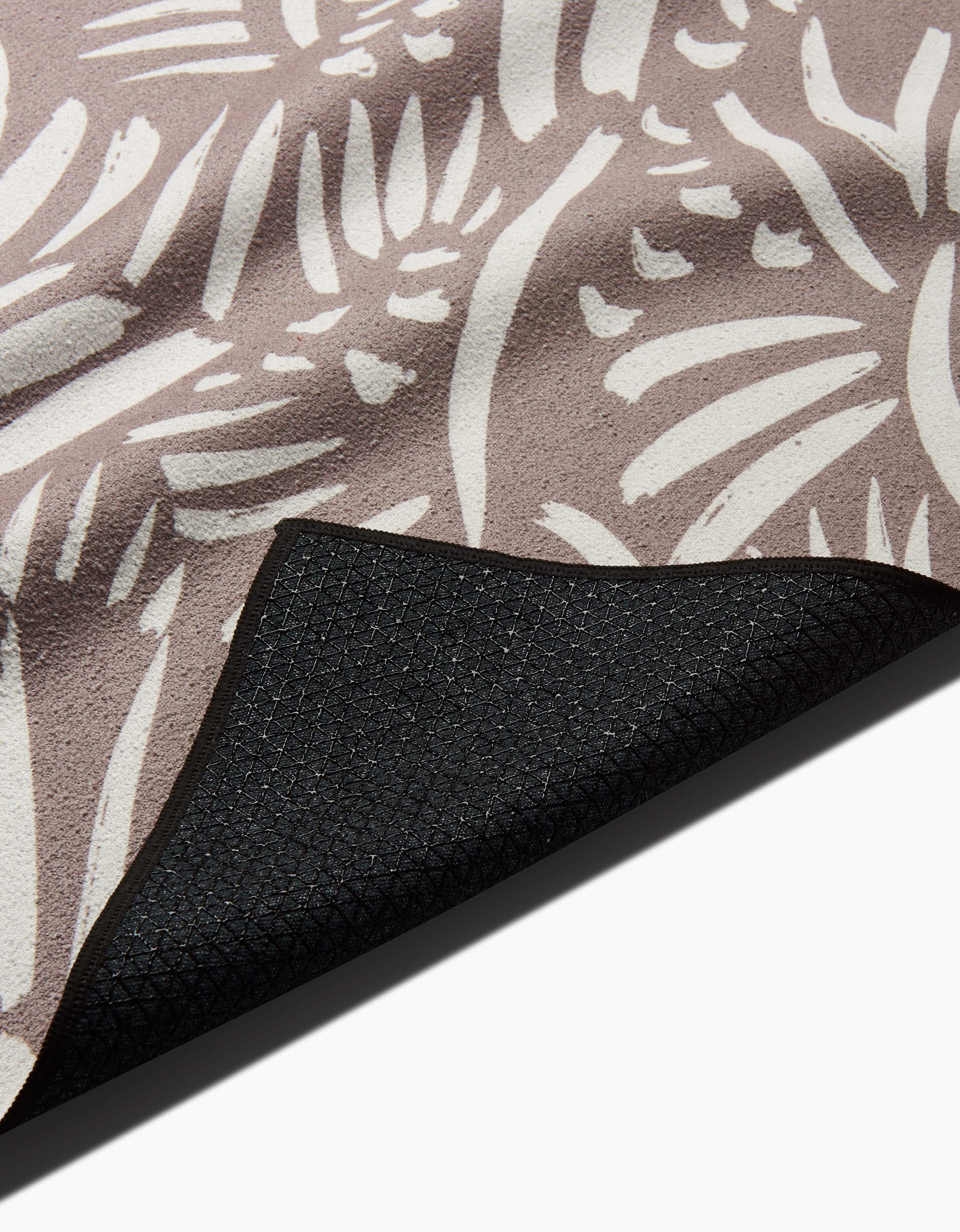 A close-up of the Miami yoga mat by Hufton Studio shows its beige surface with a white abstract floral pattern and textured black non-slip towel backing, one corner folded to reveal both sides.