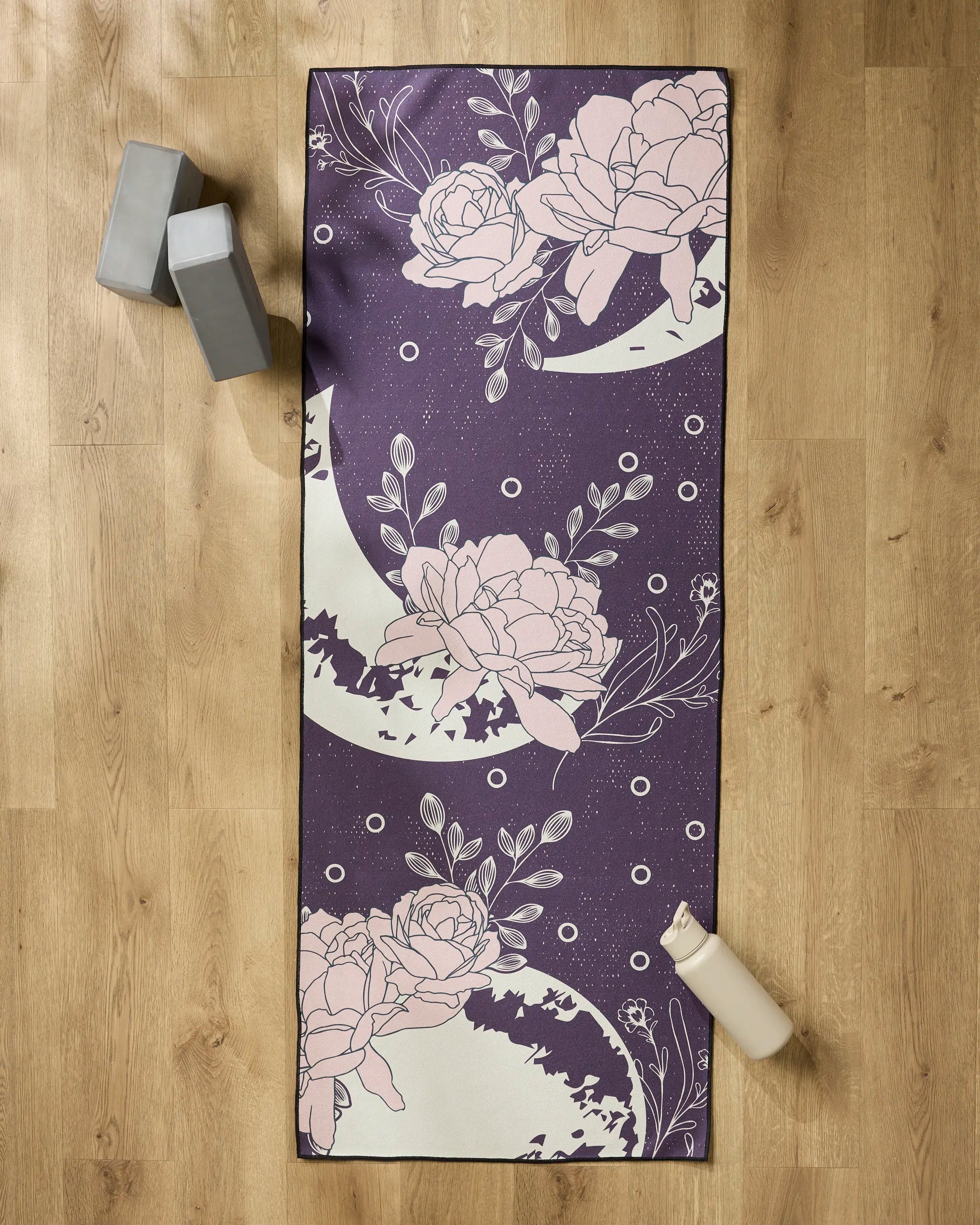 The Ashley Celena "Midnight Garden" yoga mat, featuring a purple background with bold pink florals, is spread on a wooden floor and topped with a non-slip towel. Two gray blocks and a white water bottle complete the inviting setup.