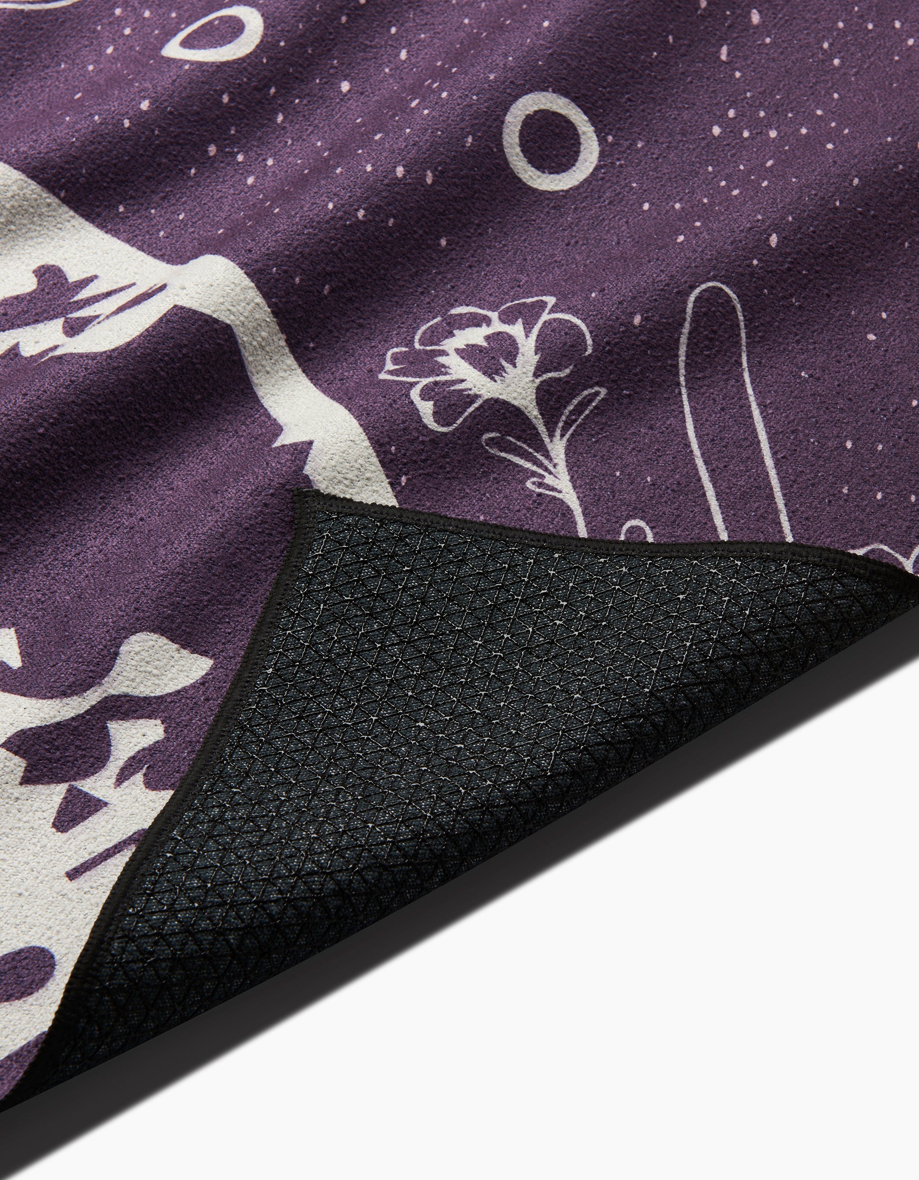 A close-up of the Ashley Celena Midnight Garden yoga towel, showcasing purple fabric with white floral and abstract patterns and a curled textured black slip-resistant backing.