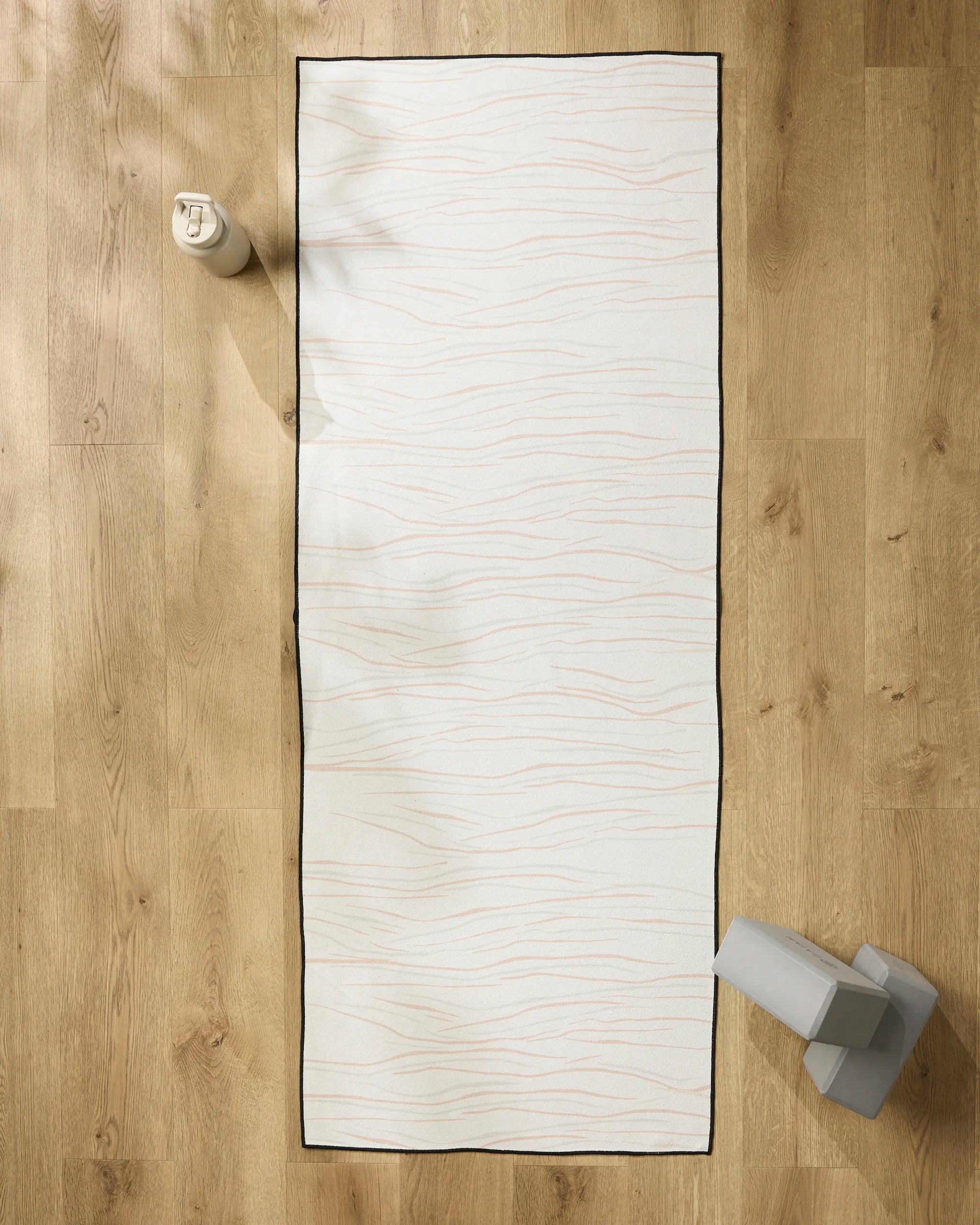 The GEOMETRY Mind white yoga mat with thin wavy lines and a black border lies on a light wooden floor, topped with a non-slip towel. A water bottle sits at the top left, and grey yoga blocks are placed at the bottom right.