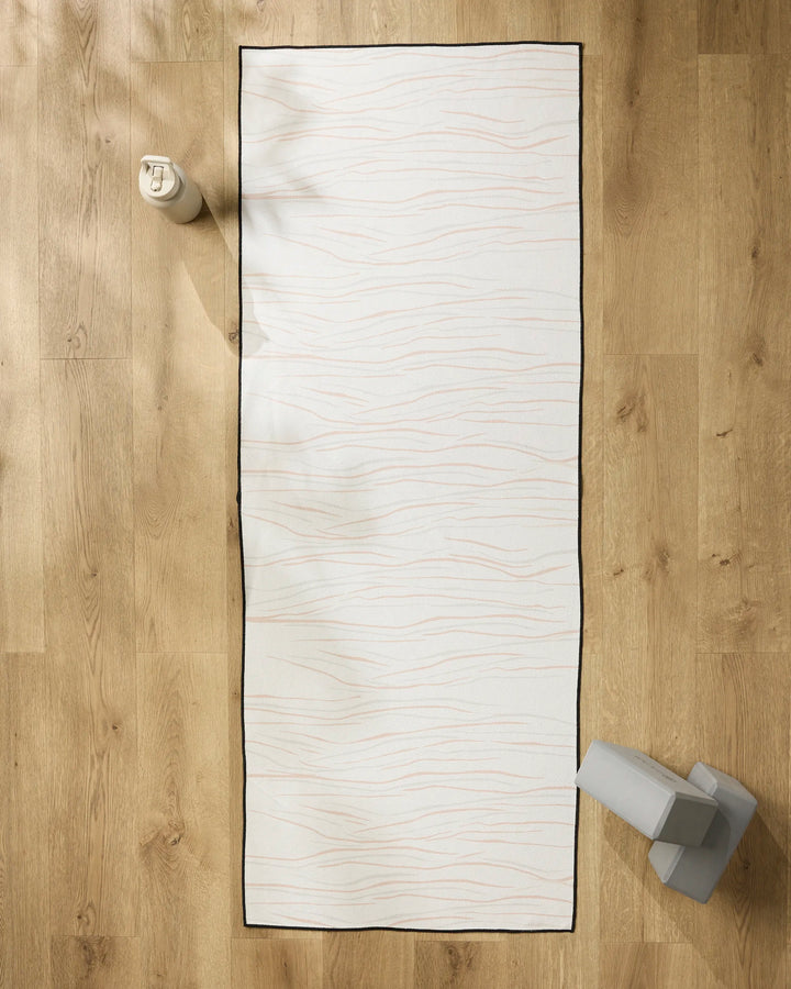 The GEOMETRY Mind white yoga mat with thin wavy lines and a black border lies on a light wooden floor, topped with a non-slip towel. A water bottle sits at the top left, and grey yoga blocks are placed at the bottom right.