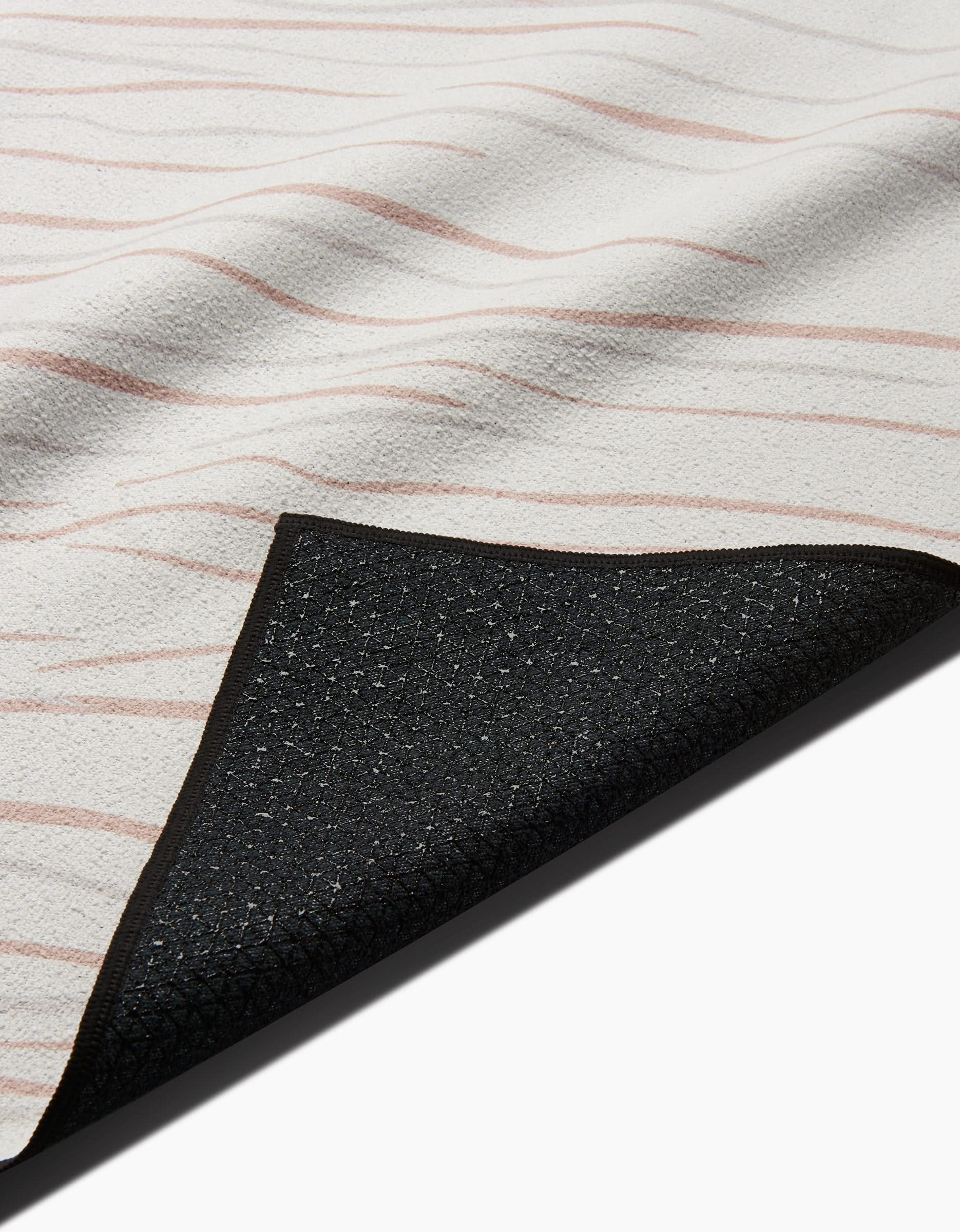 A close-up of GEOMETRYs Mind rug—a cream-colored textured surface with thin wavy pink lines—shows a corner flipped to reveal its slip-resistant black backing.