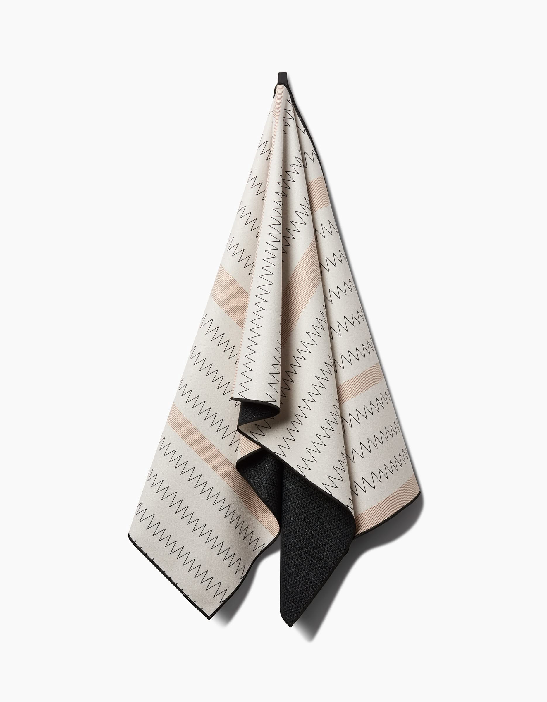 The Coit Creative Mod Zig Zag yoga towel in beige and cream features zigzag patterns and horizontal stripes. A contrasting dark gray reverse side creates a bold edge along the bottom as it hangs on a hook.