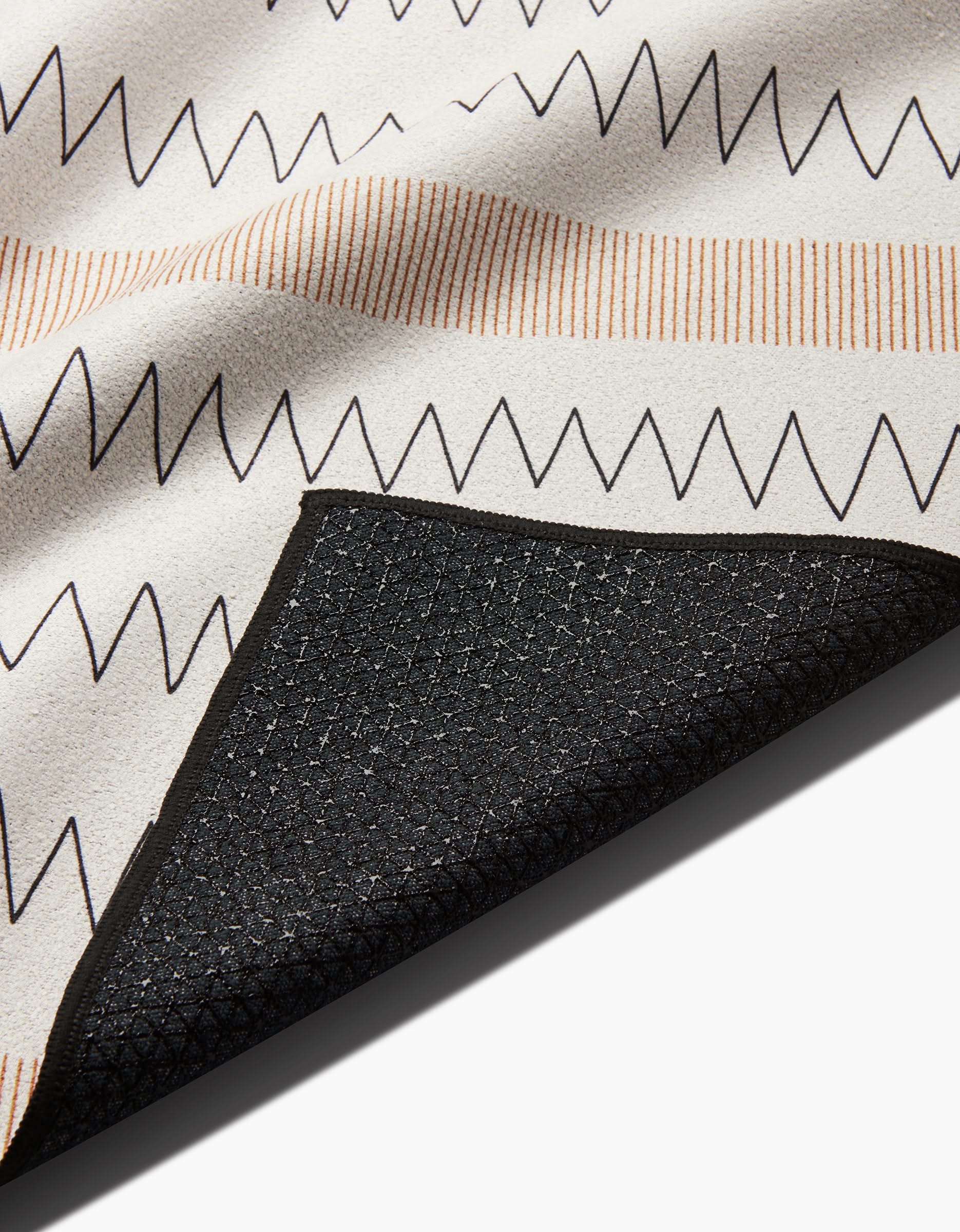 A close-up of the Coit Creative Mod Zig Zag non-slip yoga towel with black and beige zigzag stripes on white, and a folded corner showing the textured black underside.