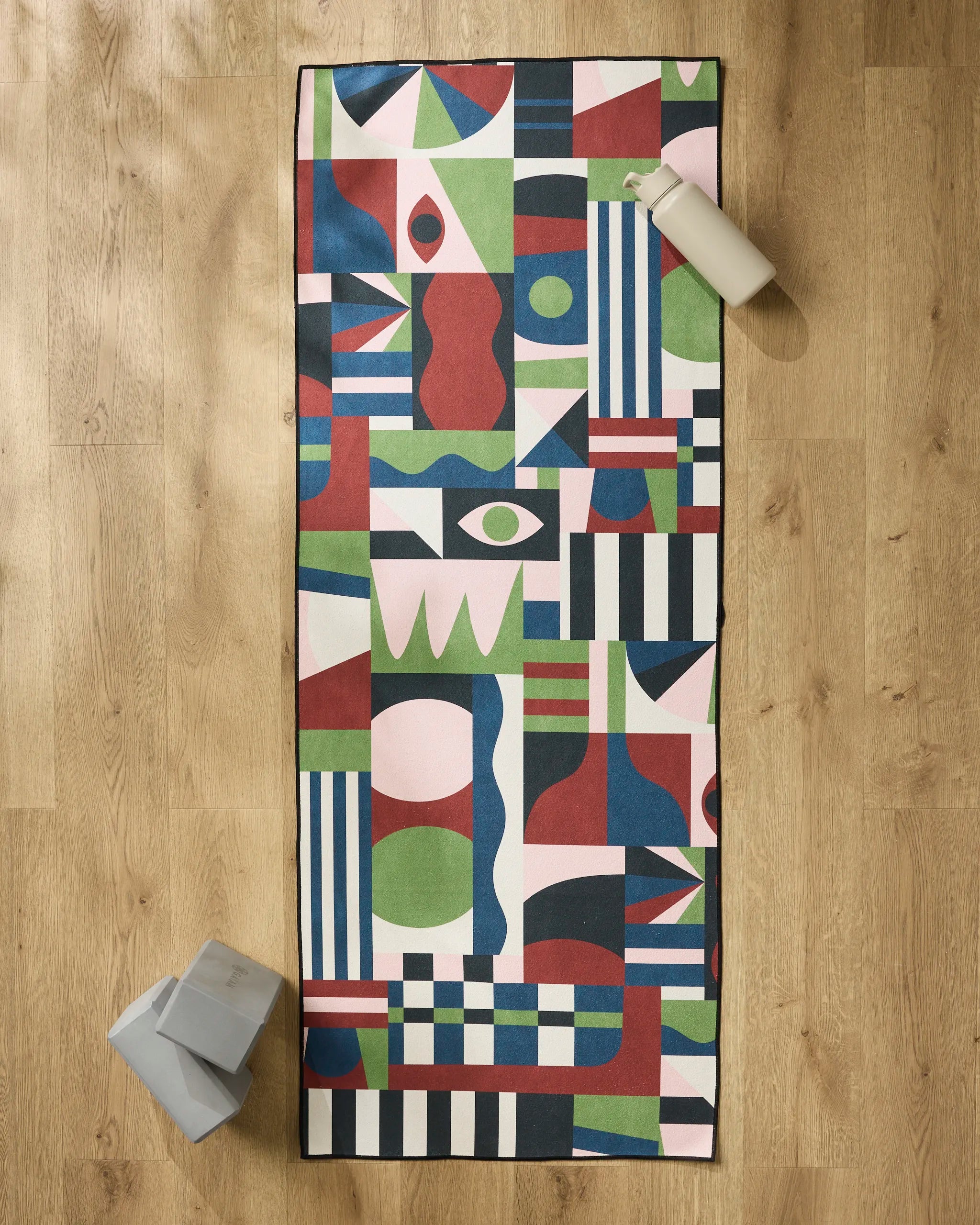 The Modern Bauhaus Mood yoga mat by Claire Iglesias, featuring a colorful abstract geometric pattern, rests on a wooden floor with a recycled towel, white water bottle, and gray yoga block ready for your practice.