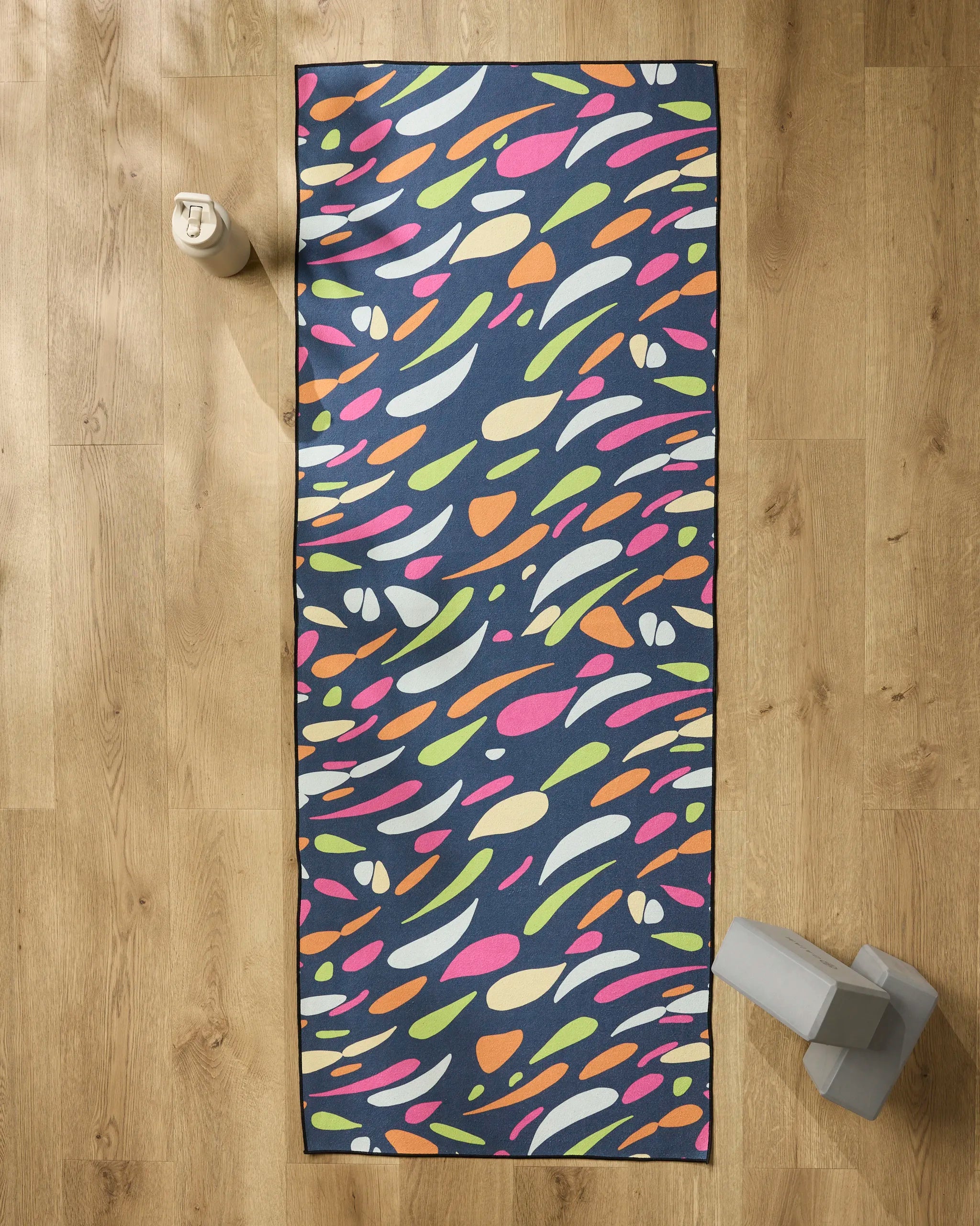The GEOMETRY Motion yoga mat features a colorful abstract pattern on a dark background. Shown on a wooden floor with a non-slip yoga towel, water bottle, and gray yoga block.
