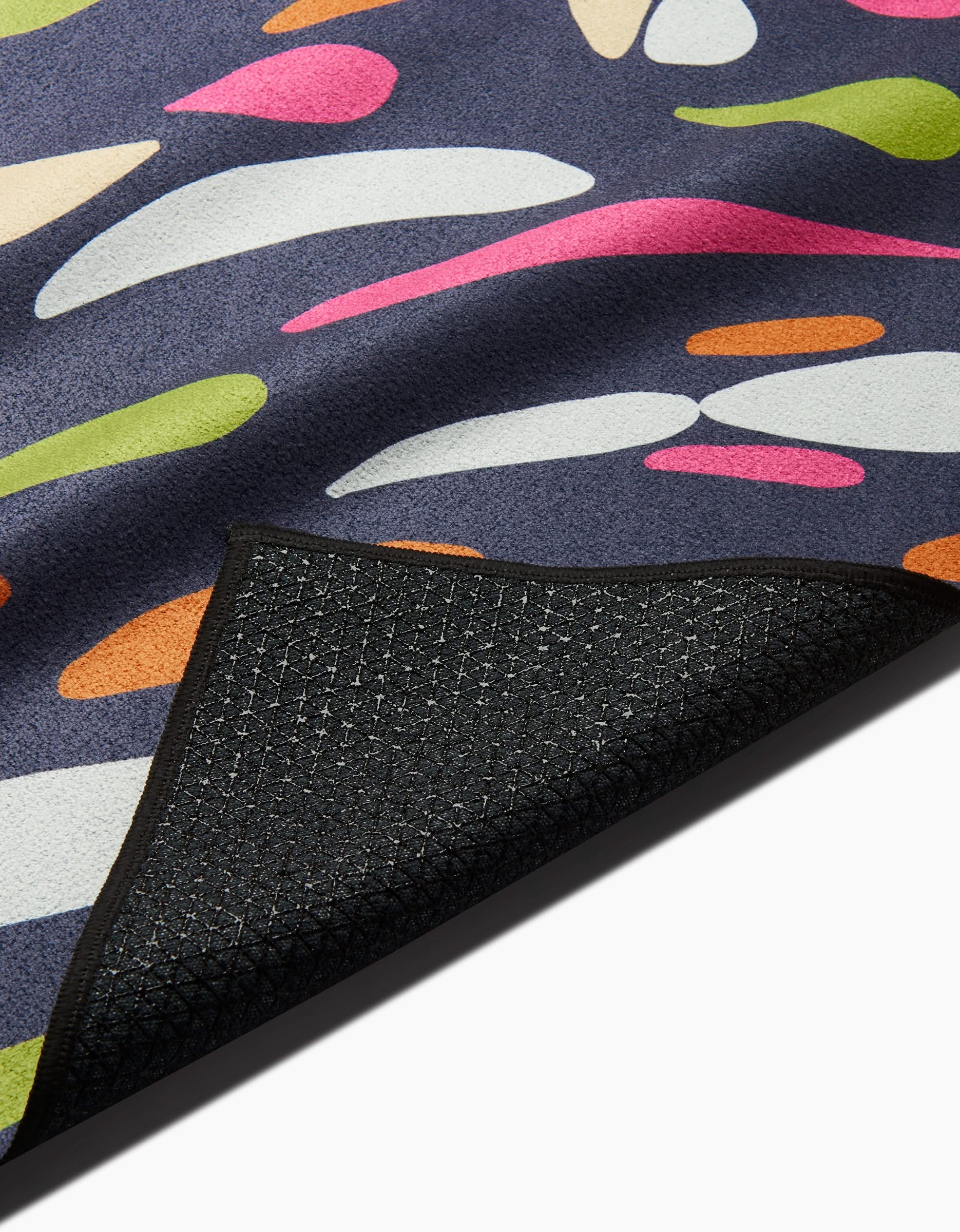 The GEOMETRY Motion rug features colorful abstract shapes in pink, green, orange, white, and cream on a dark background. A flipped corner reveals its slip-resistant backing for added security.