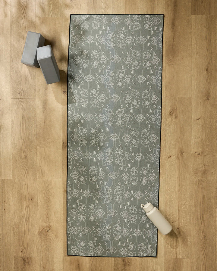 The Ash Cascade "Mountain Bloom" patterned gray yoga mat lies on a wooden floor, with two gray yoga blocks nearby, a white water bottle at the corner, and a non-slip towel on top for extra grip.