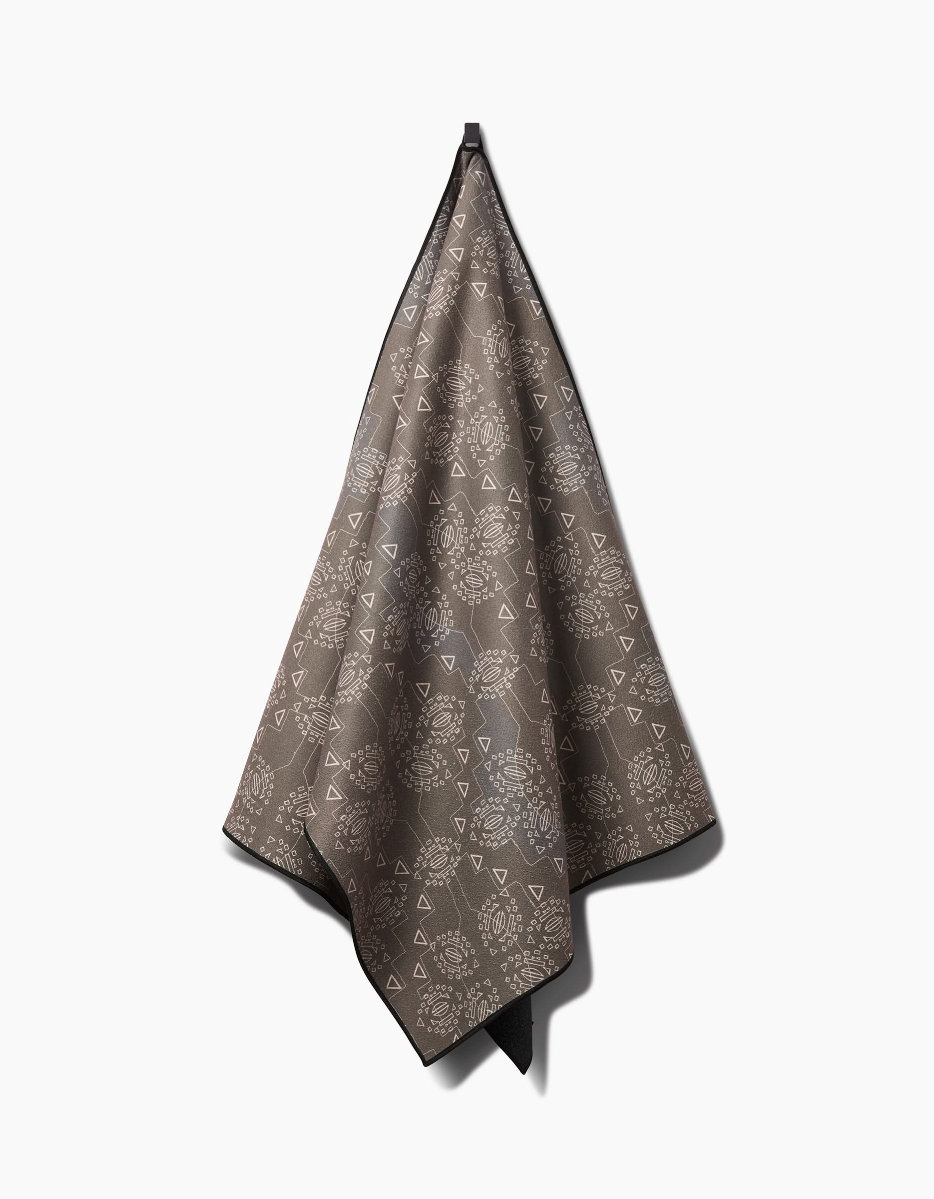 The Ash Cascade Mountain Bloom is a brown yoga towel with a hanging loop, decorated with white geometric and circular patterns. Shown on a plain white background, it’s ideal as a recycled, eco-friendly yoga towel.