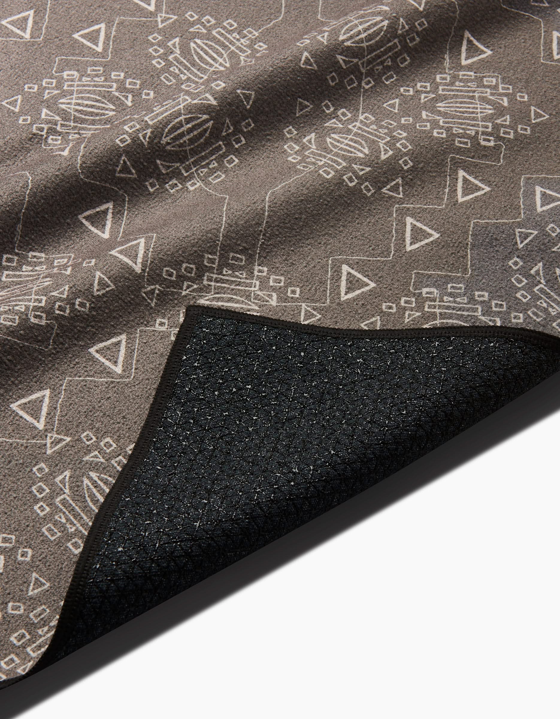 A close-up of the Ash Cascade Mountain Bloom mat with geometric brown and beige patterns, featuring a corner flipped to reveal its black textured, non-slip backing for added stability, similar to a silicone grip yoga towel.