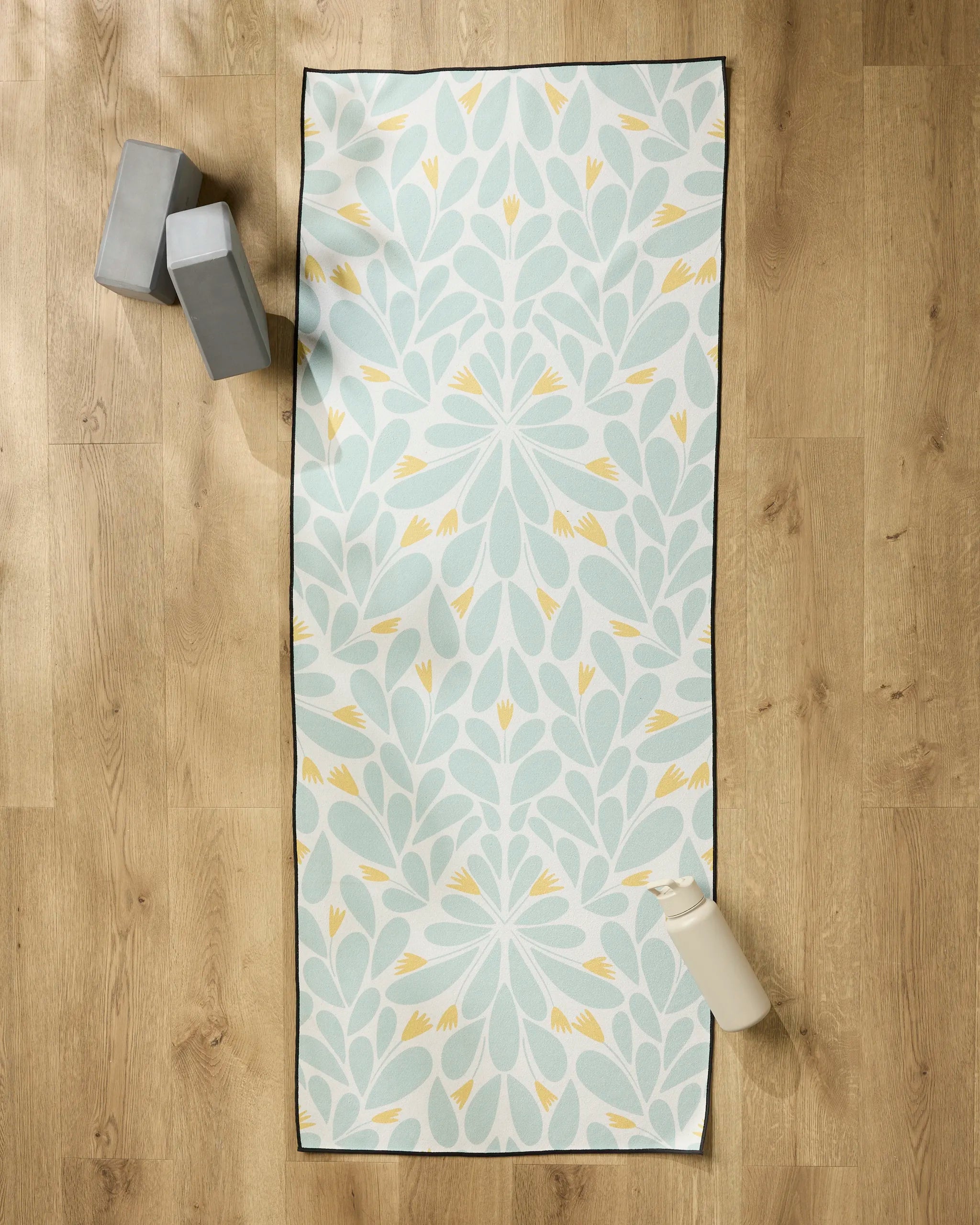 The Rebecca Woolbright Mustard Pinwheel yoga mat with a floral design is laid out on a wooden floor, topped with a non-slip towel. Two gray yoga blocks and a white water bottle are placed nearby.