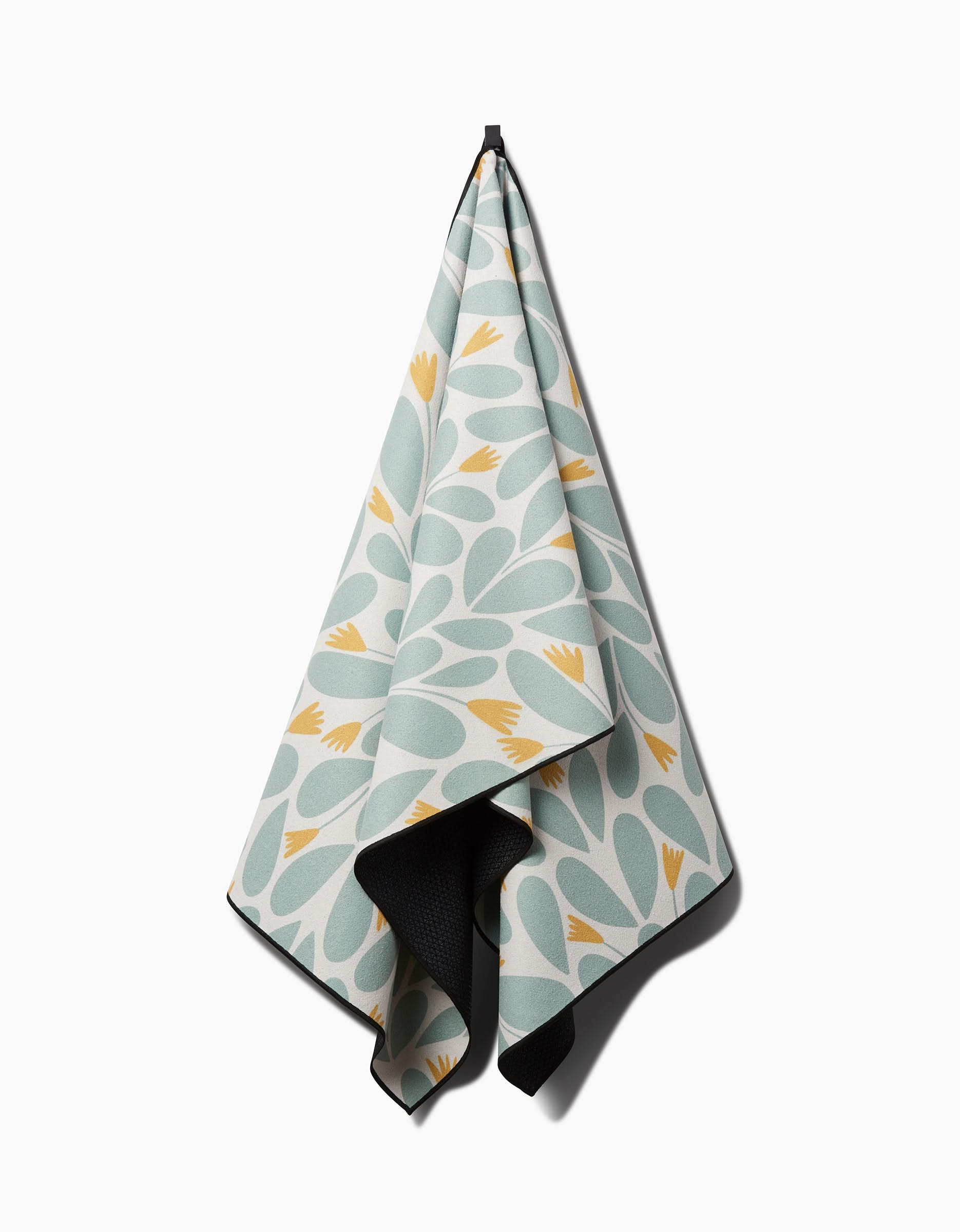 The Rebecca Woolbright Mustard Pinwheel yoga towel, featuring light blue and yellow leaf-like patterns and made from recycled materials, hangs on a hook against a plain white background.