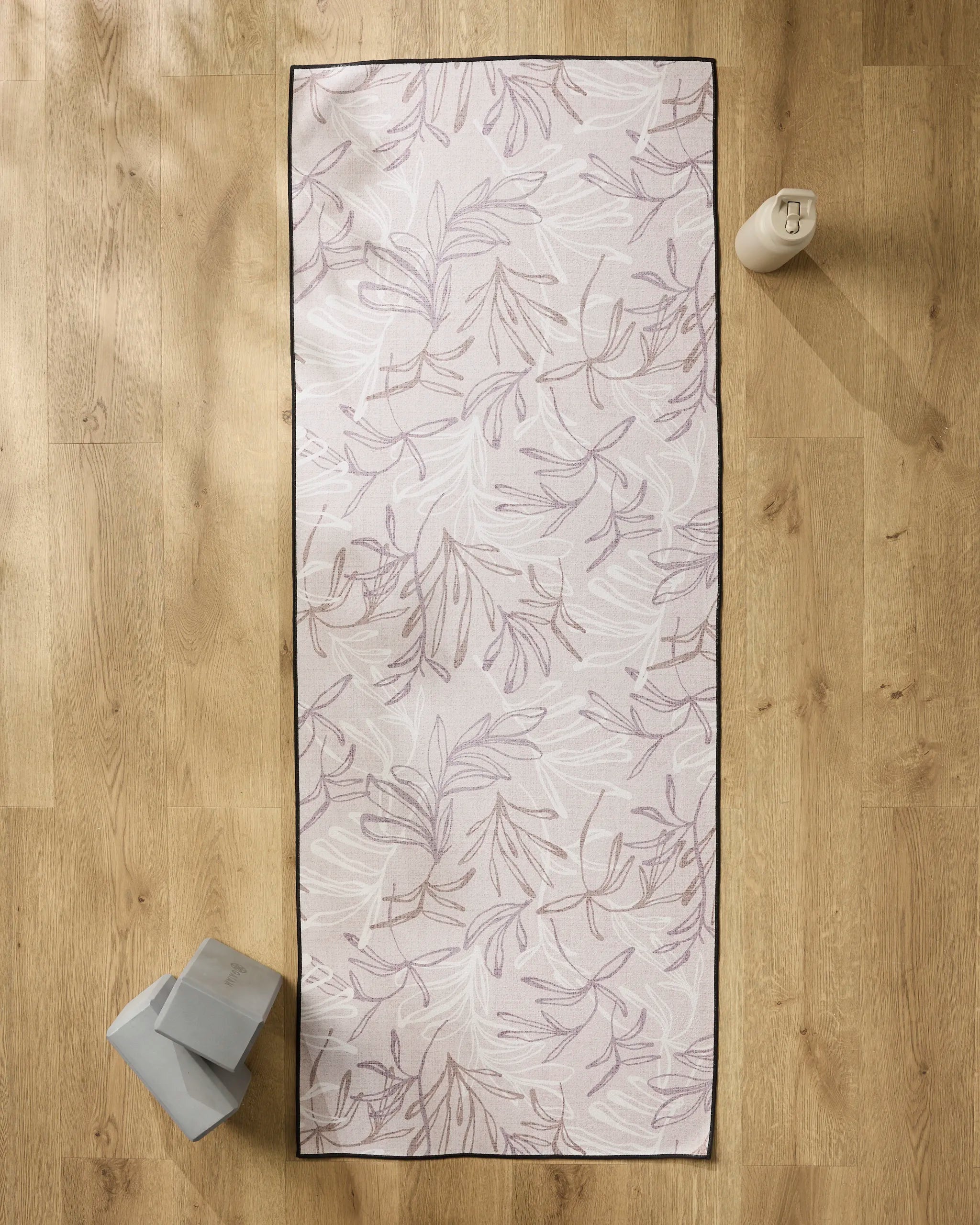 The Kris Ruff Muted Meadow yoga mat with a subtle leaf pattern rests on a wooden floor, topped with a non-slip towel. A water bottle sits at the top right, while two yoga blocks are placed at the bottom left.
