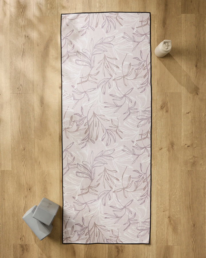 The Kris Ruff Muted Meadow yoga mat with a subtle leaf pattern rests on a wooden floor, topped with a non-slip towel. A water bottle sits at the top right, while two yoga blocks are placed at the bottom left.