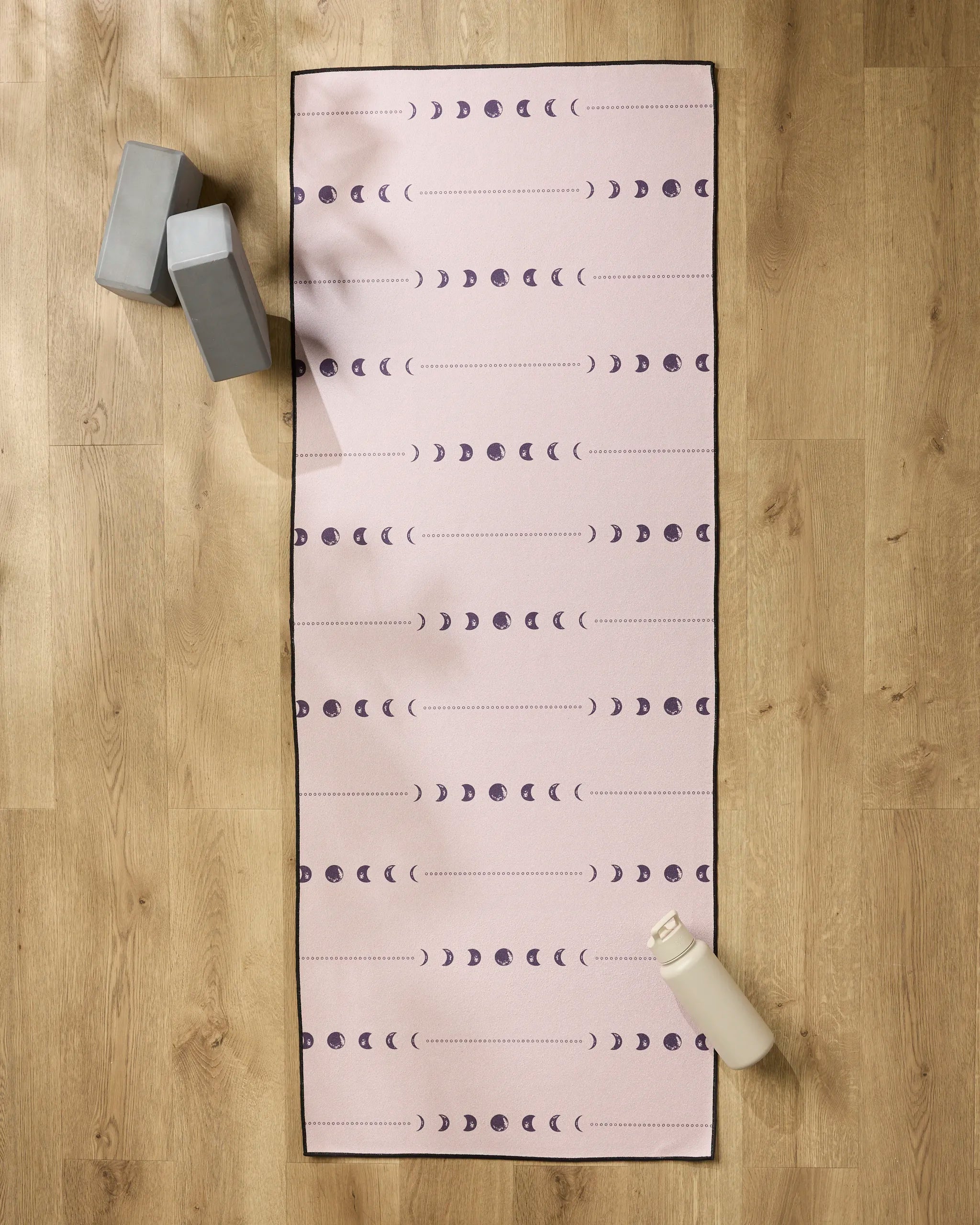 The "Night Sky" yoga mat by Ashley Celena, featuring purple moon phases on pink with a black border, lies on a wooden floor with a non-slip towel. Nearby are two gray yoga blocks and a white water bottle.