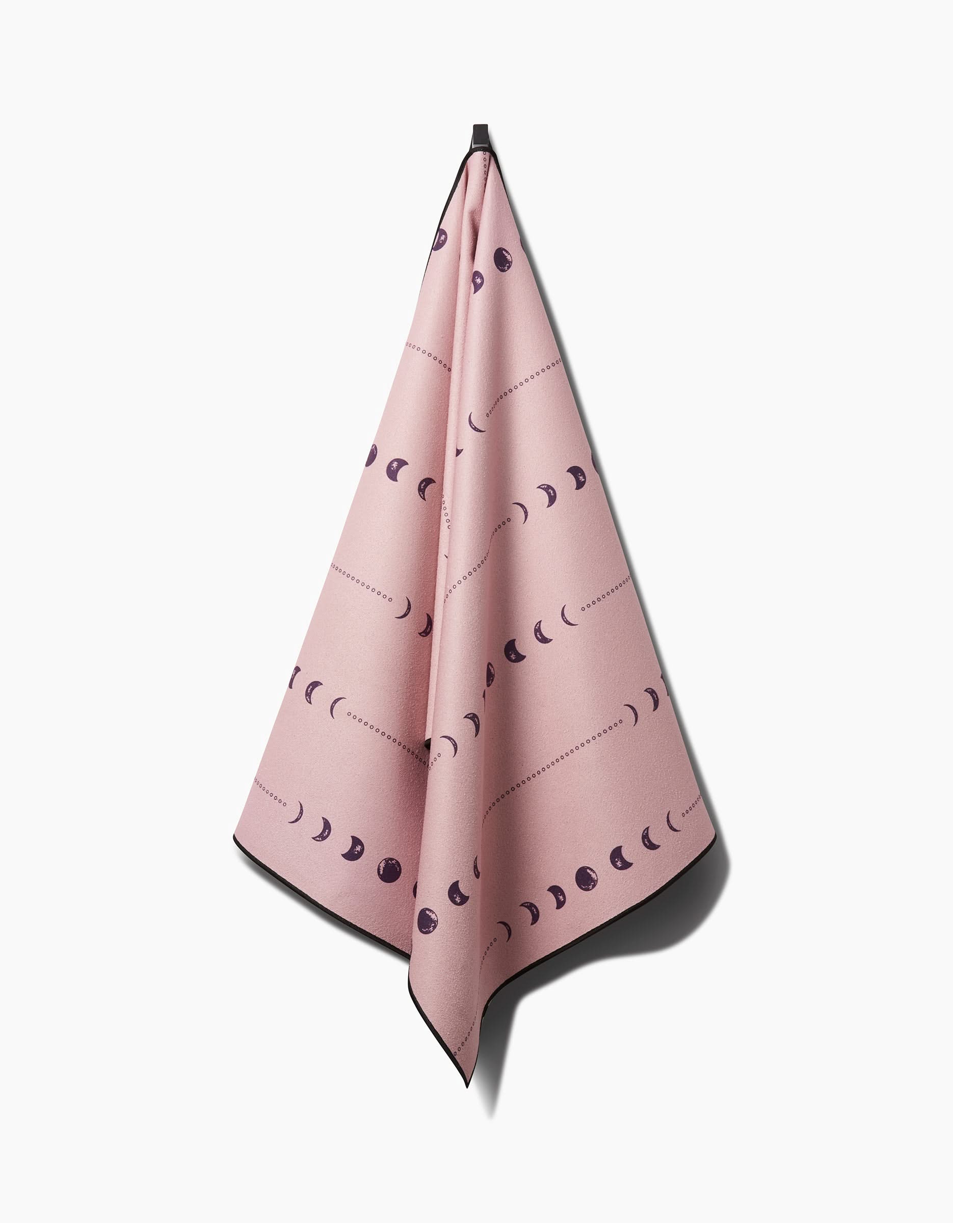 The Ashley Celena Night Sky pink silicone grip yoga towel, featuring black moon phases, hangs from a single hook against a white background.