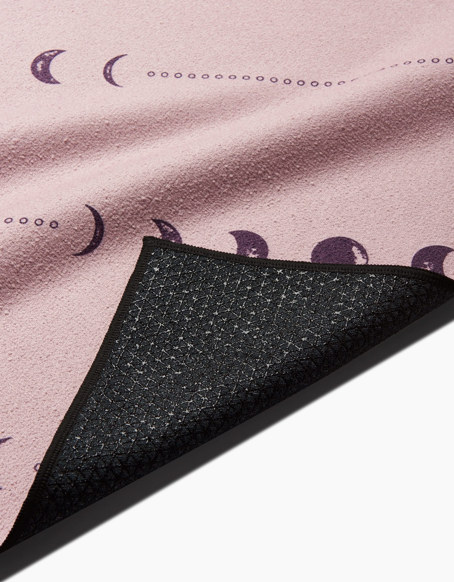 A close-up of the Ashley Celena Night Sky mat shows its pink surface with purple moon phases. A flipped corner reveals a black, silicone grip yoga towel underside featuring a geometric pattern for superior traction.
