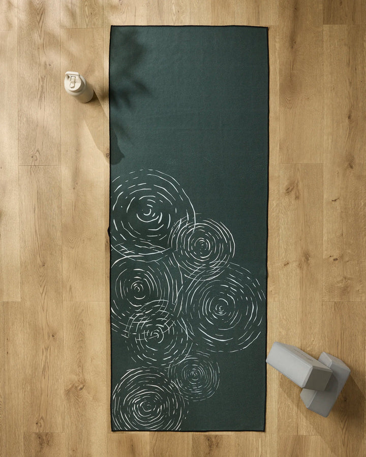 The Om yoga mat by GEOMETRY, featuring dark green color with white swirl patterns, is spread on a light wood floor. A water bottle sits at the top left and a folded non-slip towel or block is at the bottom right corner.