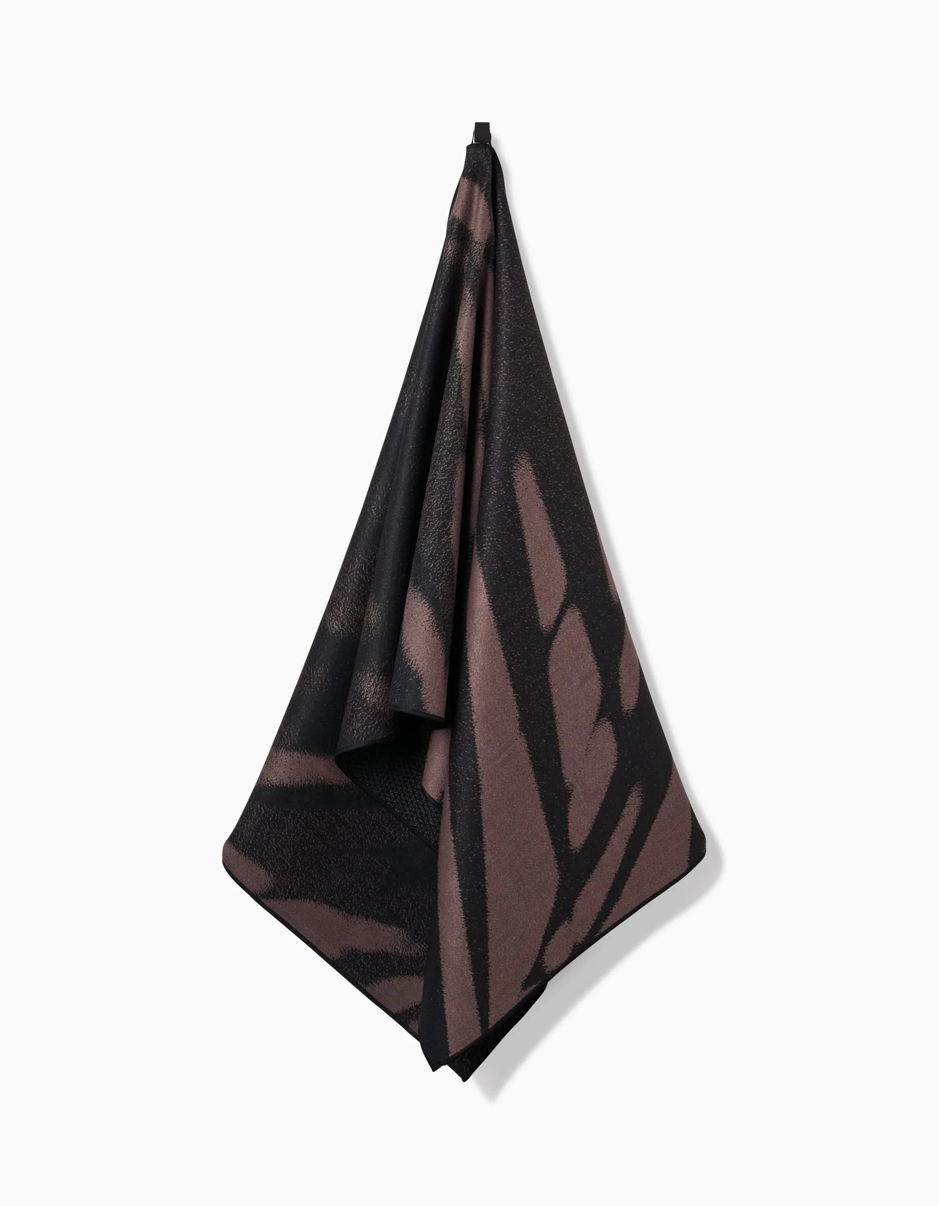 The Palm Shadow yoga towel by GEOMETRY, featuring a dark brown and black pattern and crafted from recycled materials, hangs from a hook against a white background.