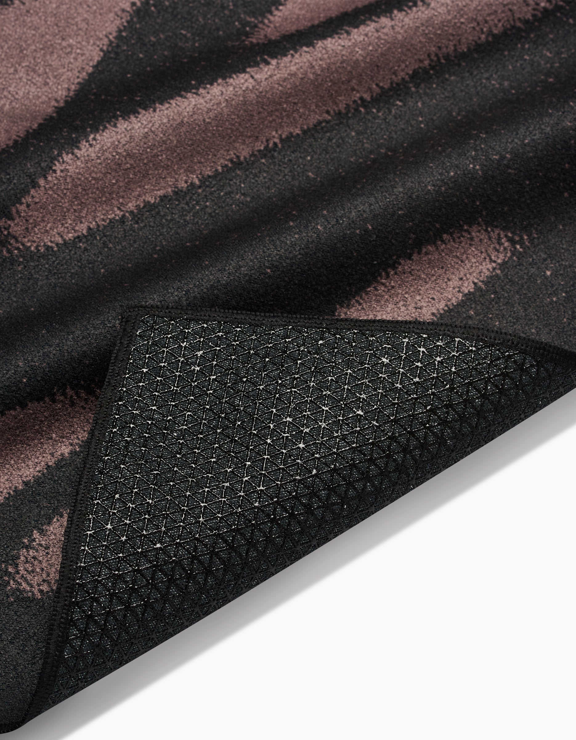 A close-up of GEOMETRYs Palm Shadow rug corner, flipped to reveal its black textured slip-resistant backing. The surface displays black and muted mauve brush strokes, photographed against a white background.