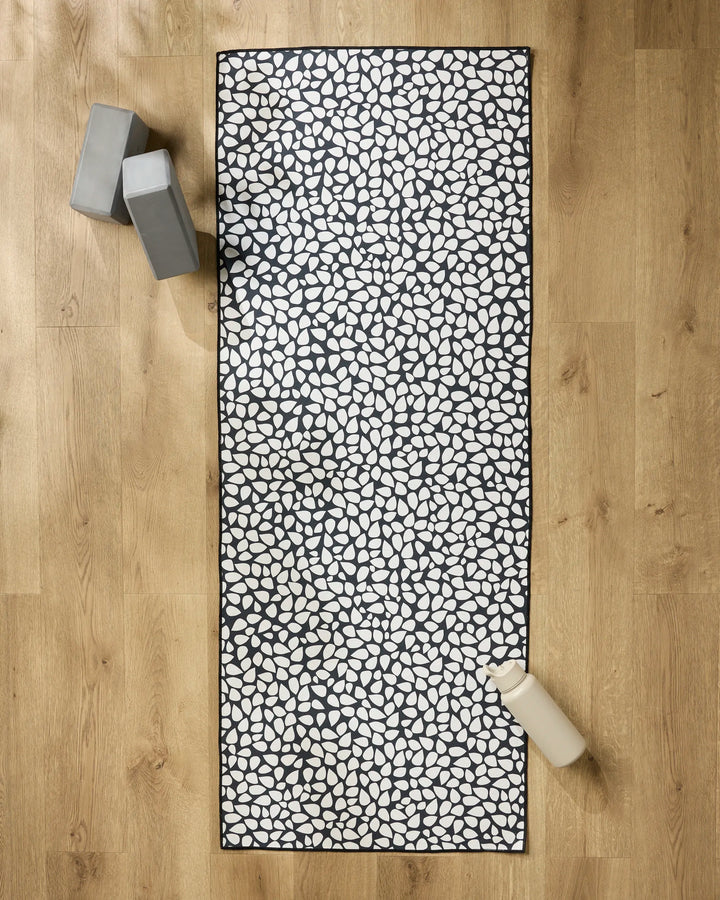The Pebbles yoga mat by Marina Solodka, featuring a black and white pebble pattern, lies on a wooden floor with a non-slip towel. Two gray yoga blocks are at the top left, and a beige water bottle sits at the bottom right.
