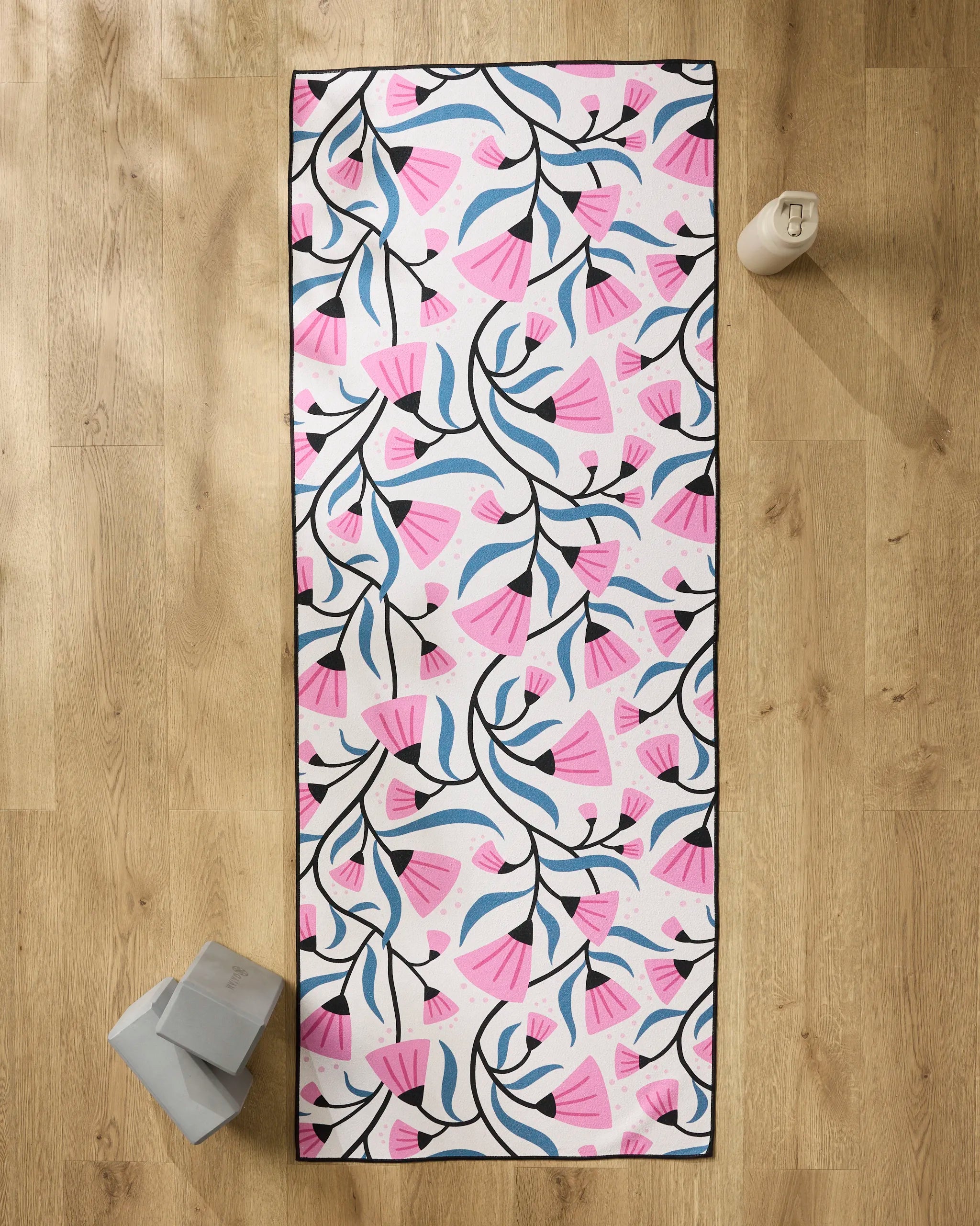 The Anette Heiberg Petal Symphony yoga mat, featuring a pink, blue, and black floral pattern, lies on a wooden floor with a reusable water bottle, two gray yoga blocks, and a non-slip yoga towel nearby.