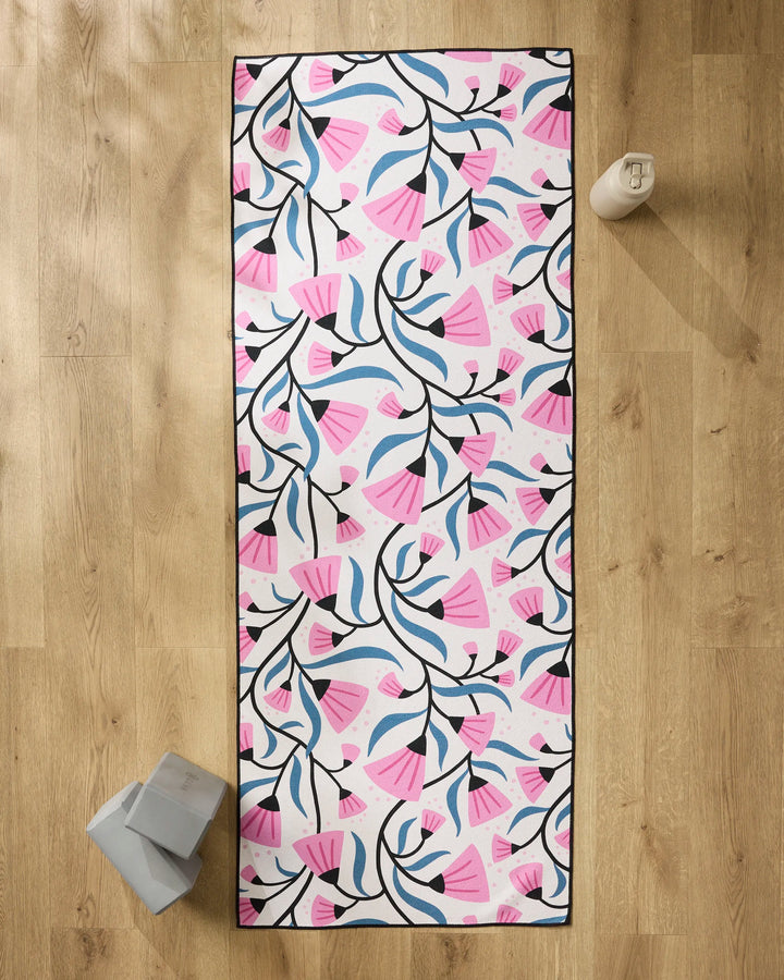 The Anette Heiberg Petal Symphony yoga mat, featuring a pink, blue, and black floral pattern, lies on a wooden floor with a reusable water bottle, two gray yoga blocks, and a non-slip yoga towel nearby.