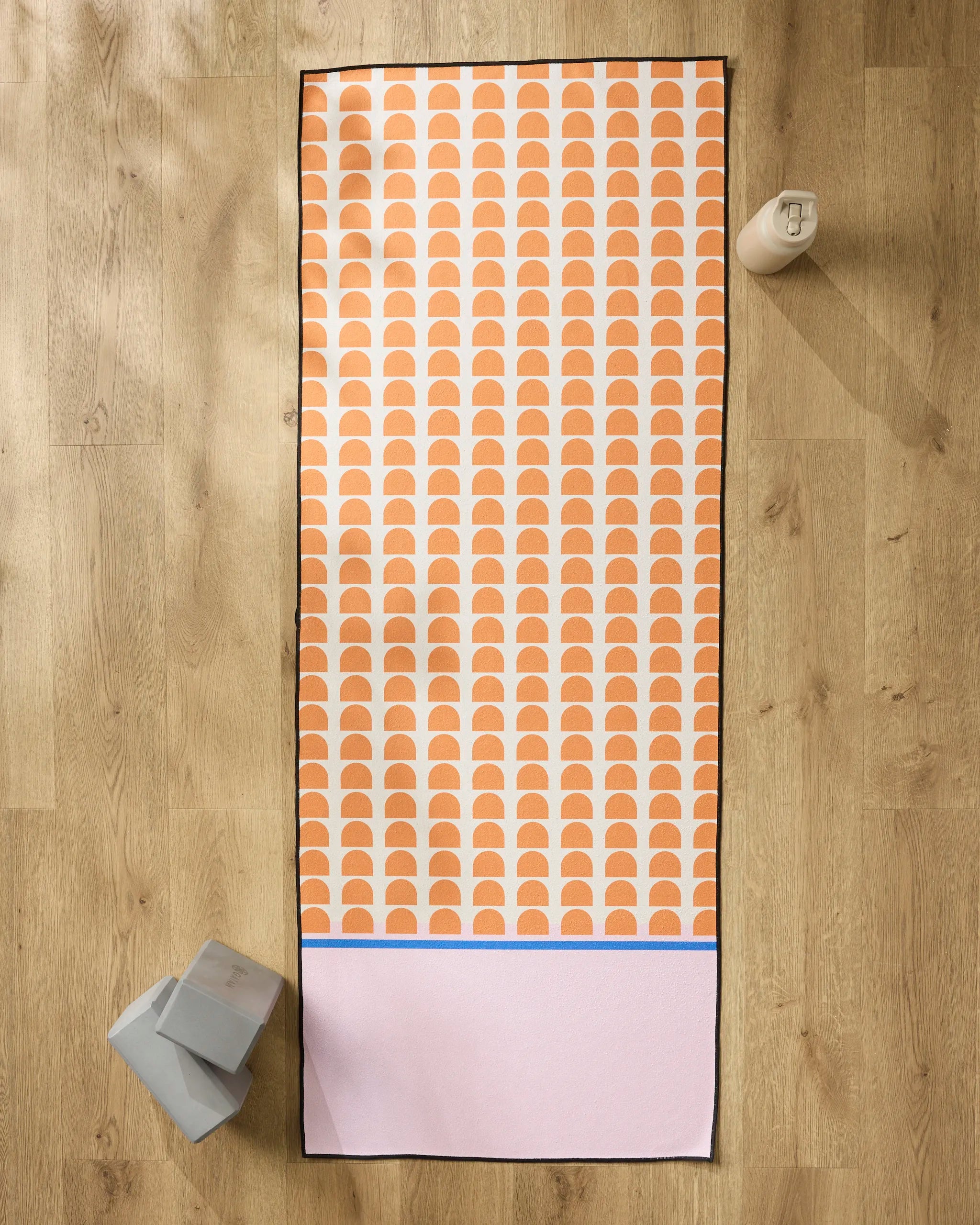 The Happy Hemera Pink Illusion yoga mat with an orange and white semi-circle design rests on a wooden floor, two gray yoga blocks to the left, a water bottle to the right, and a non-slip towel placed close by.