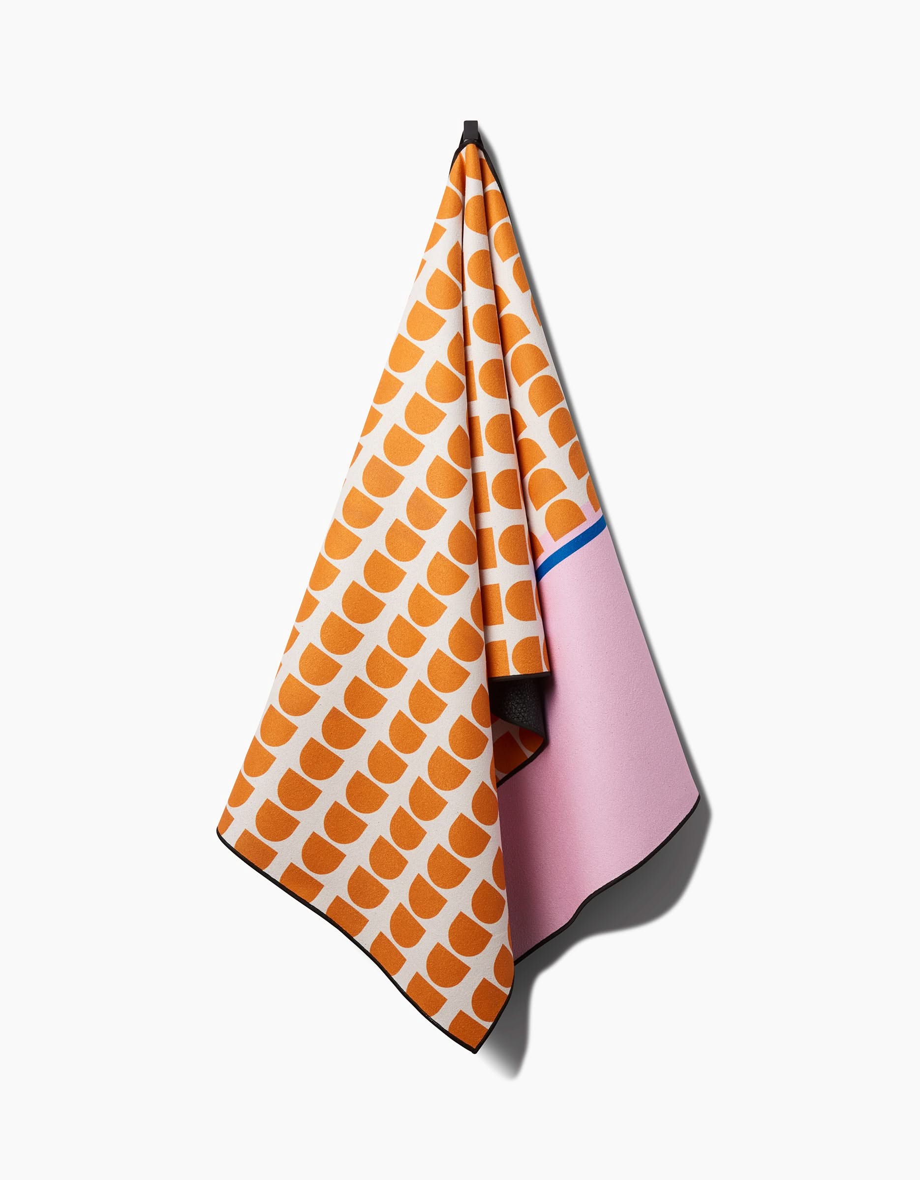 The Happy Hemera Pink Illusion yoga towel, made from recycled materials, hangs on a hook with an orange and white semicircle pattern on one side and solid pink with a blue stripe on the other, set against a plain white background.