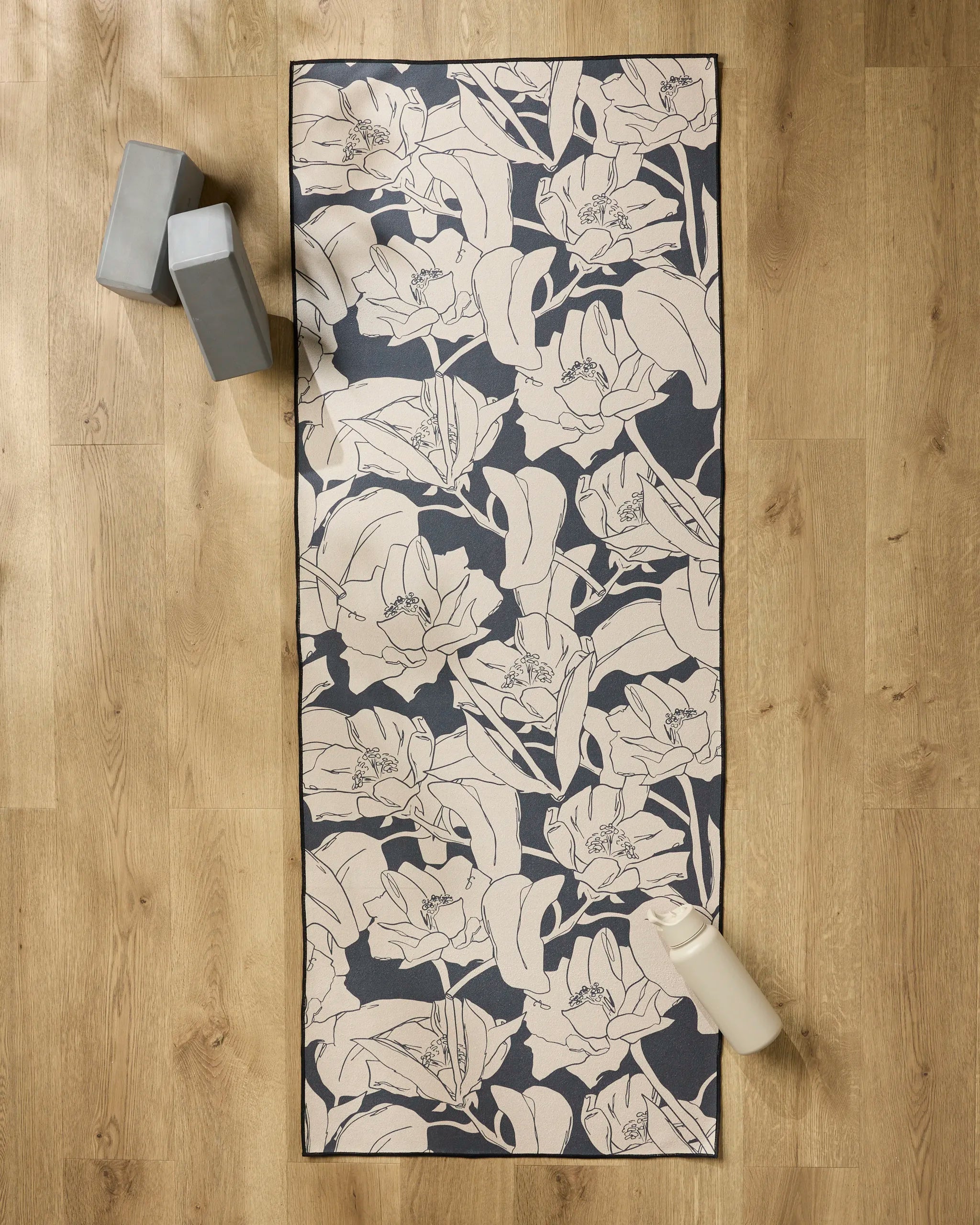 The "Plant One on Me" yoga mat by Aubrey Fairchild rests on a wooden floor, topped with a silicone grip towel for stability, while two gray yoga blocks and a white water bottle are placed nearby.