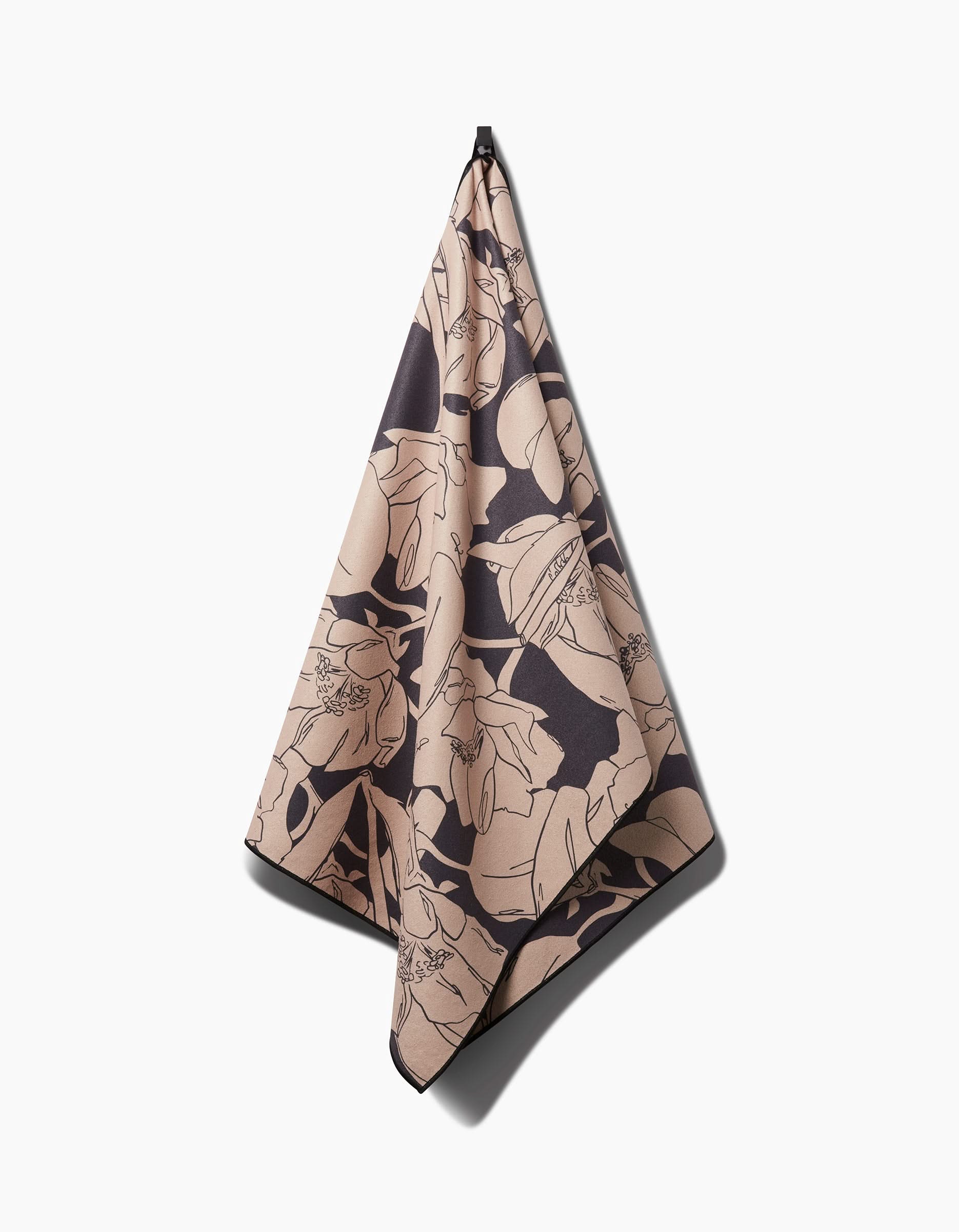 The Plant One on Me yoga towel by Aubrey Fairchild, featuring a large dark floral design, hangs on a hook against a plain white background.