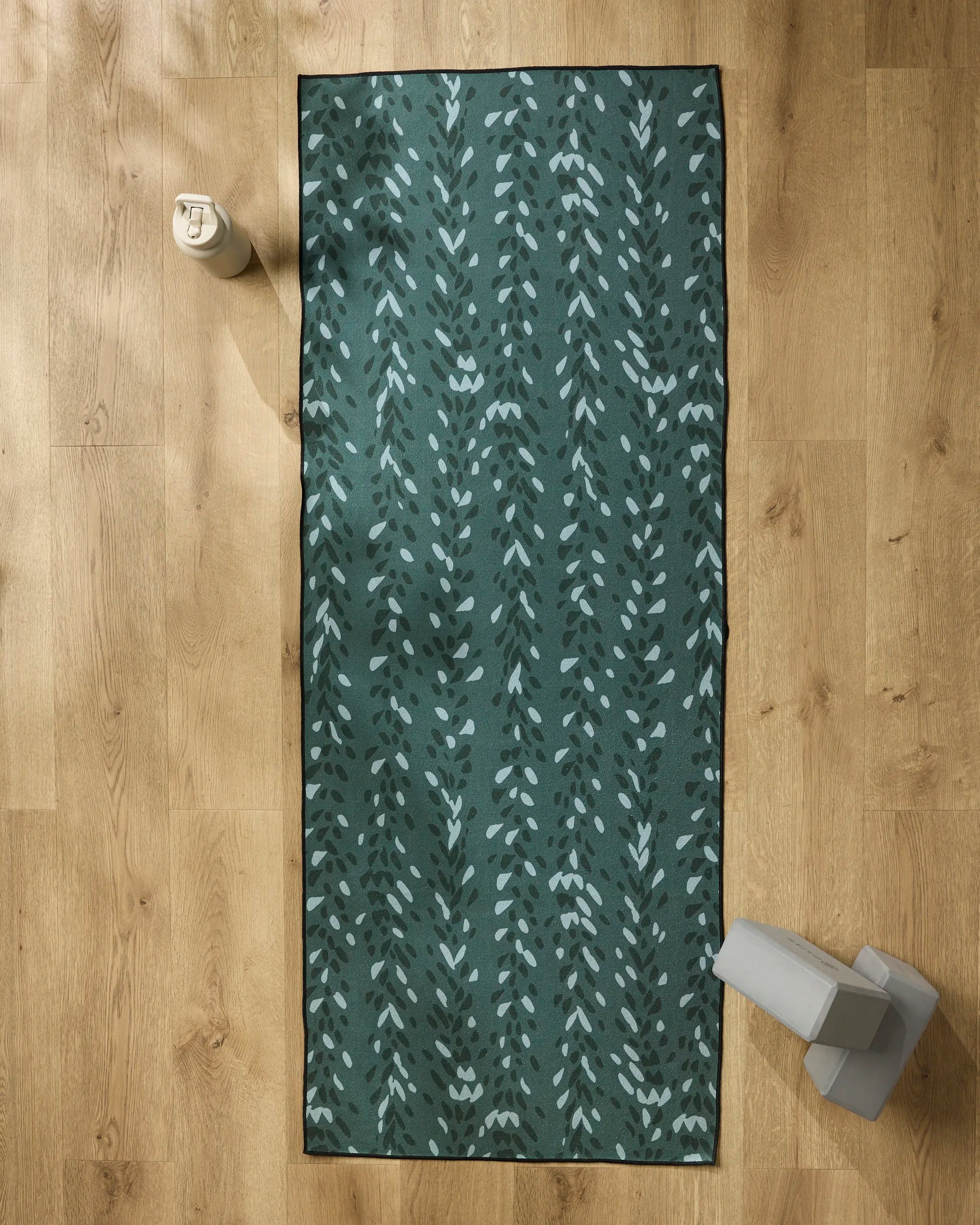 The GEOMETRY Pose yoga mat with green leaf patterns is spread on a wooden floor, featuring a non-slip towel. A water bottle is at the top left, and light gray dumbbells are placed at the bottom right.