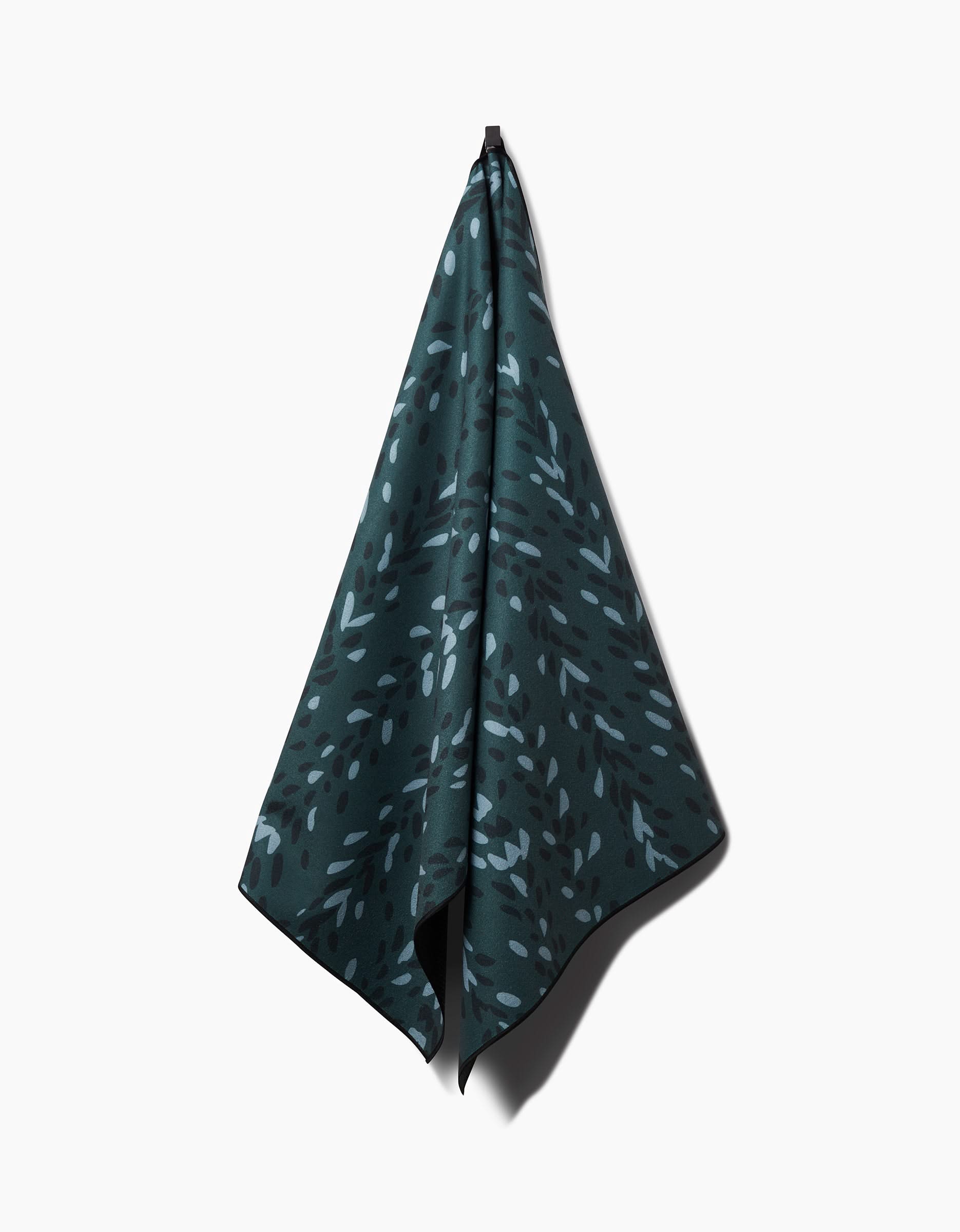 The GEOMETRY Pose yoga towel, crafted from recycled materials in dark green, displays a subtle light green abstract leafy pattern and hangs from a hook against a plain white background.