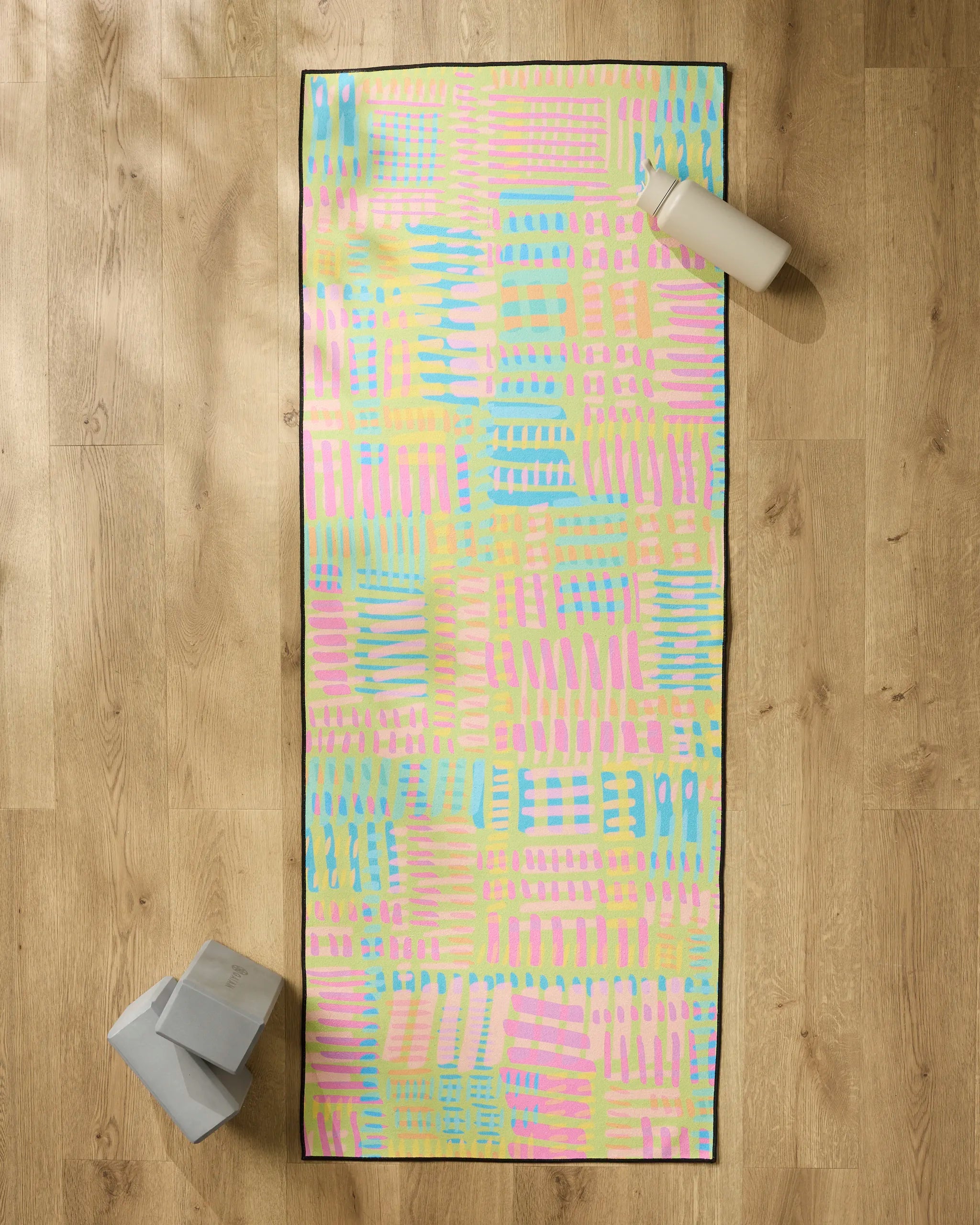 The "Positive Vibes" yoga mat by Sareka Unique, featuring a pastel abstract pattern, is laid on a wooden floor with a non-slip towel. Two gray blocks are at the bottom left and a white bottle sits at the top right.