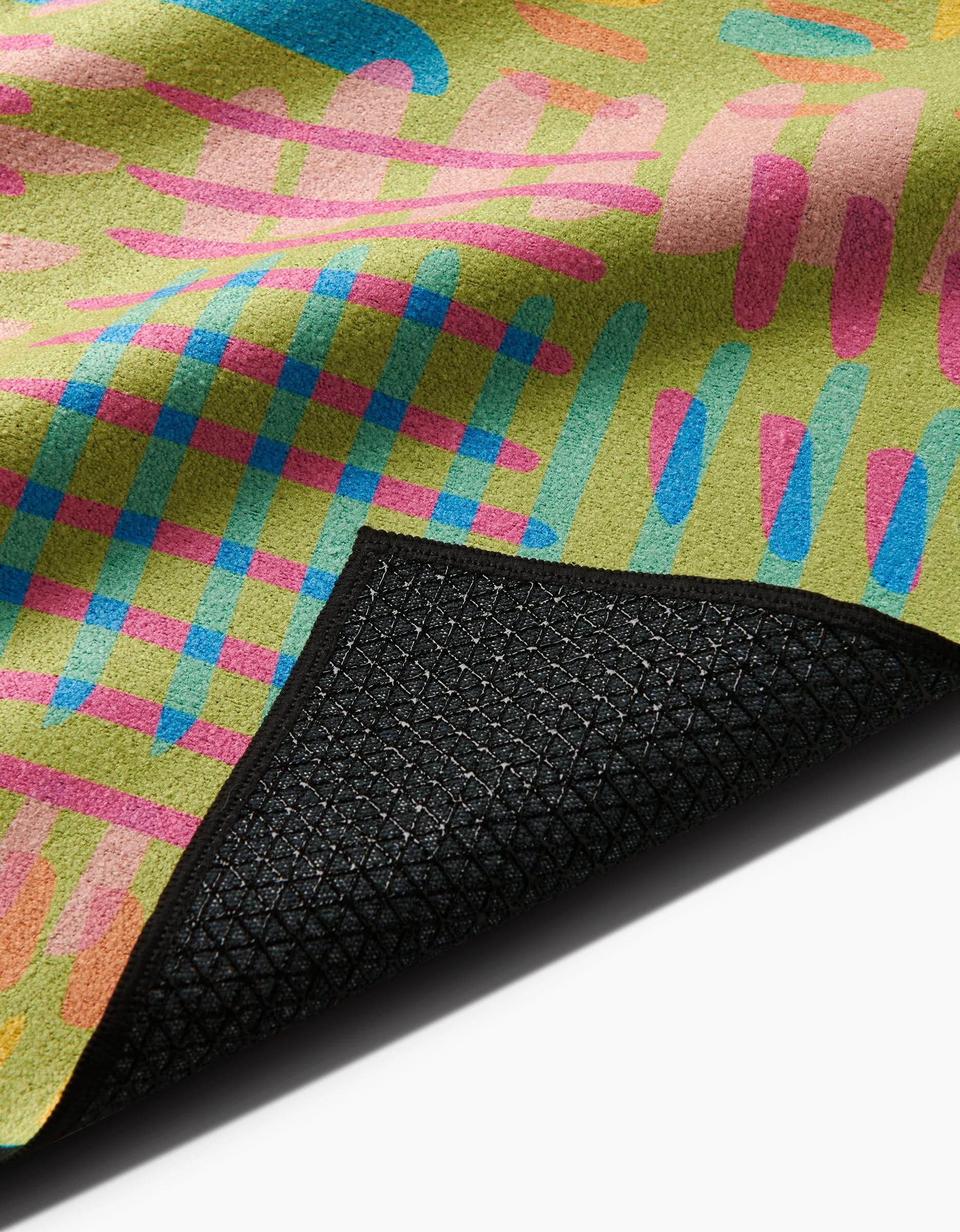 A close-up of the Sareka Unique Positive Vibes rug, featuring abstract pink, blue, and orange patterns on green; a flipped corner reveals its black textured silicone grip non-slip backing.
