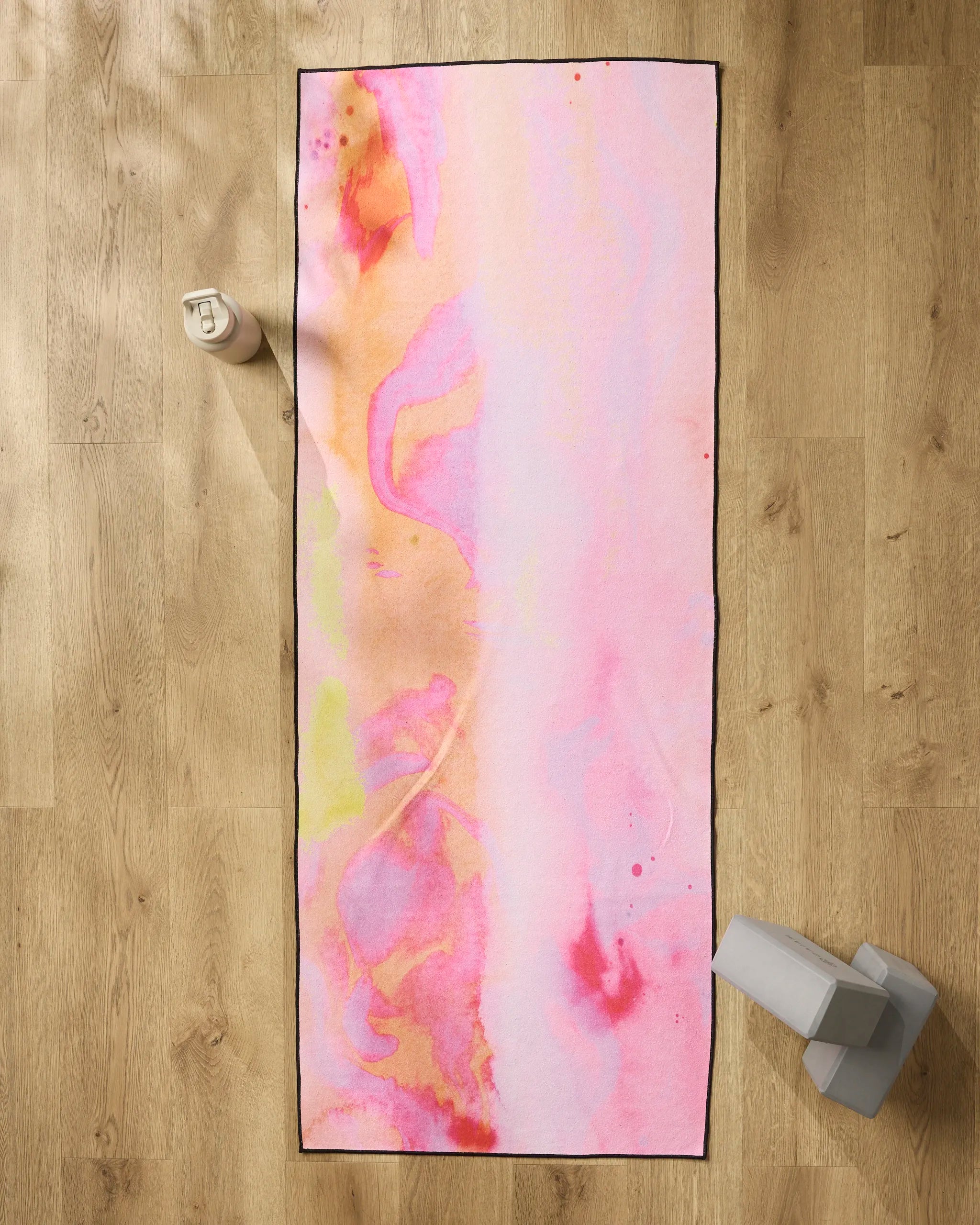The Purity yoga mat by Geometry, featuring a pink and orange abstract pattern, is laid on a wooden floor with a non-slip yoga towel, a water bottle at the top left, and two gray yoga blocks at the bottom right.