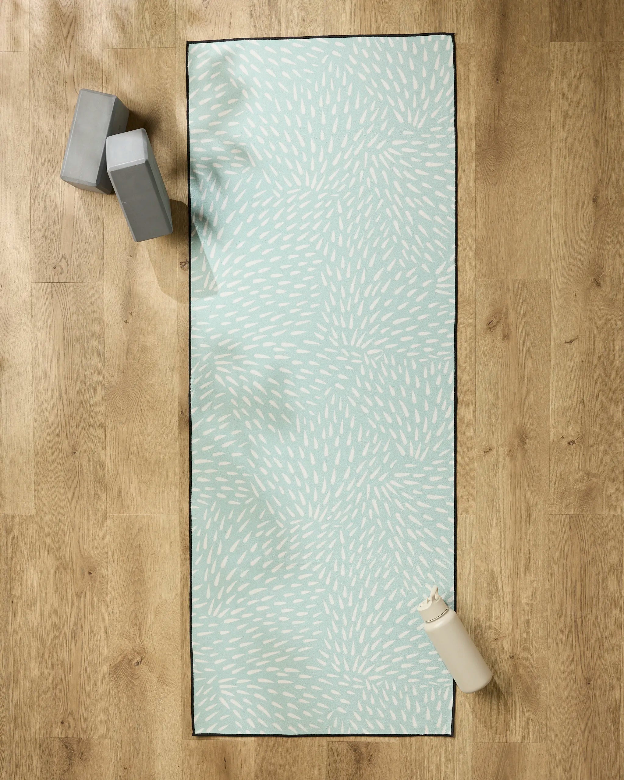 The Rebecca Woolbright Quill Blue yoga mat, featuring a white abstract pattern, is laid on a wooden floor with a non-slip towel, two gray blocks, and a white water bottle.