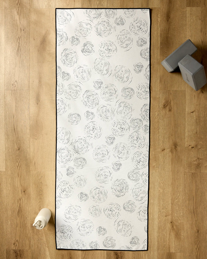 The GEOMETRY Rhythmic yoga mat, made from recycled materials and featuring white with light gray spirals, rests on a wooden floor alongside a rolled towel and two stacked yoga blocks.